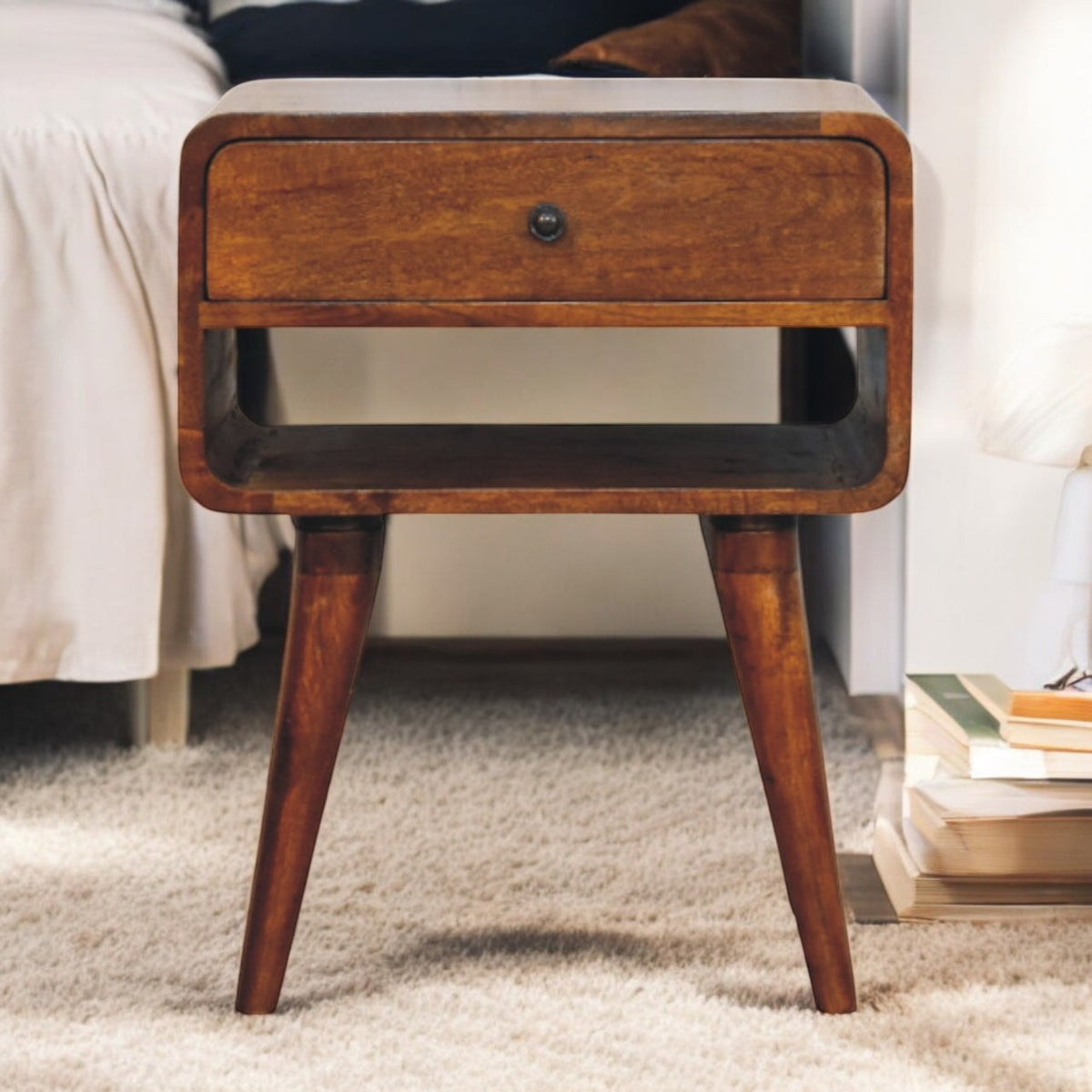 Chestnut-toned nightstand beside a bed, showing its drawer, open slot, dark knob, and tapered legs.