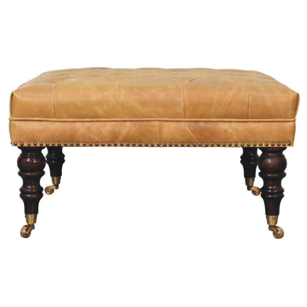 Tan upholstered ottoman with dark turned legs, brass casters, and nailhead trim, shown straight on.