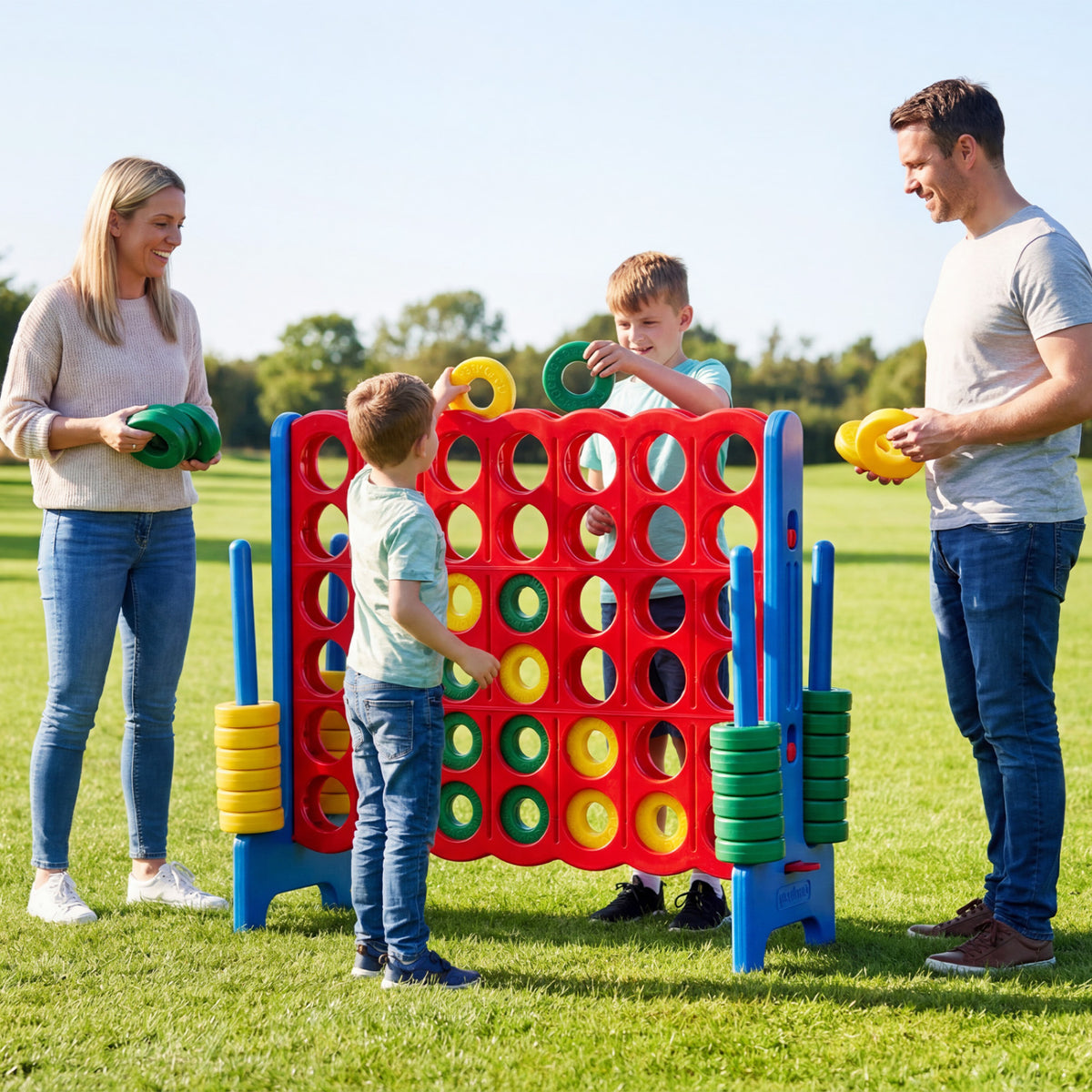 Two adults and two children play with a large red, blue, green, and yellow four-in-a-row game on a lawn.