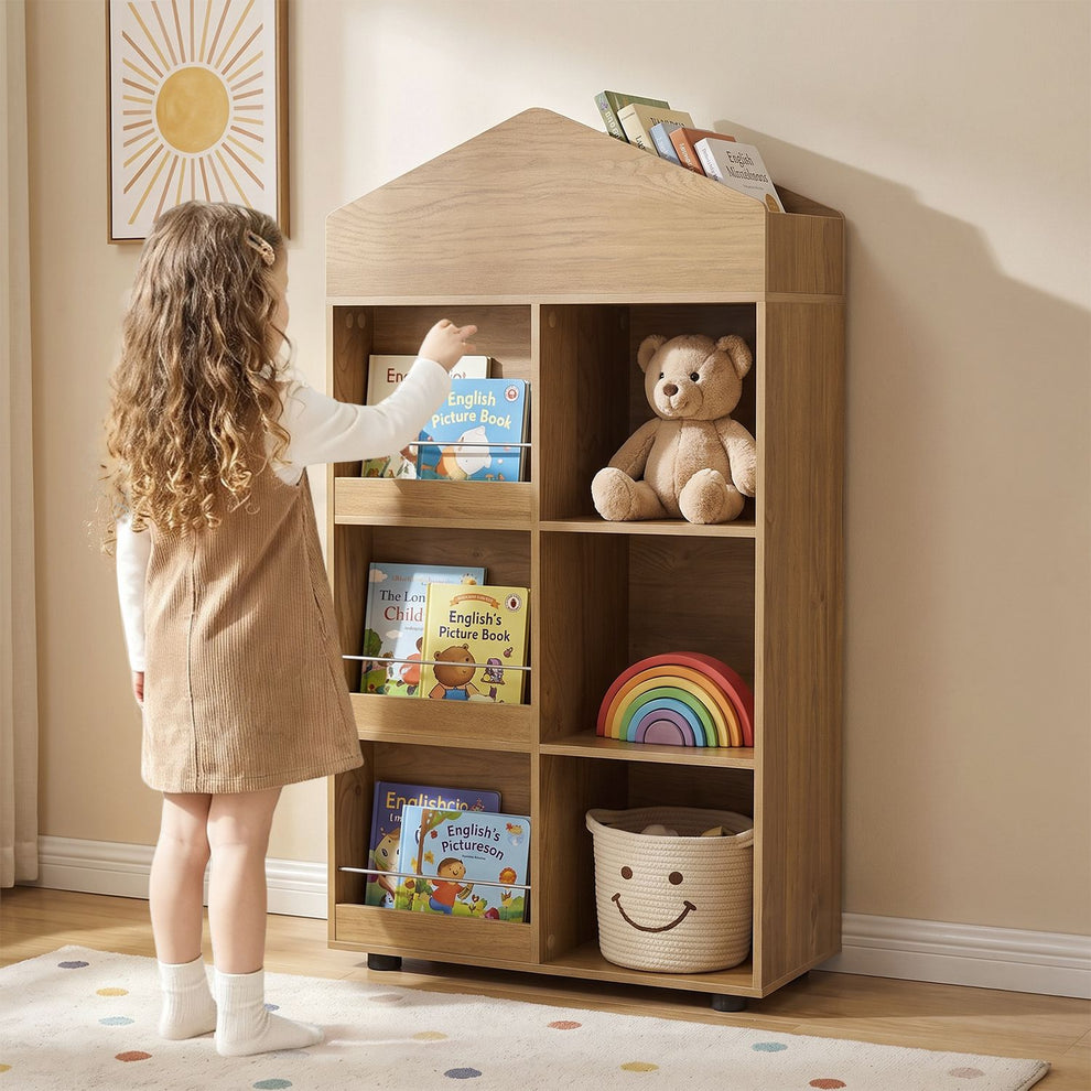 Child reaching for a book on a house-shaped wooden bookshelf filled with books, a teddy bear and toys.