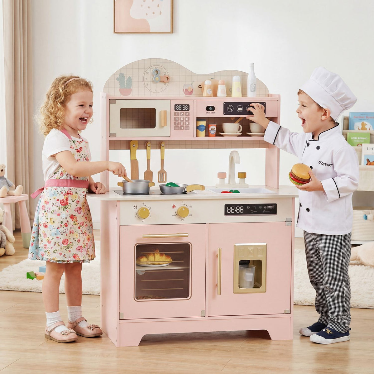 Two children play chef beside a pink toy kitchen with an oven, stovetop, sink and shelves.