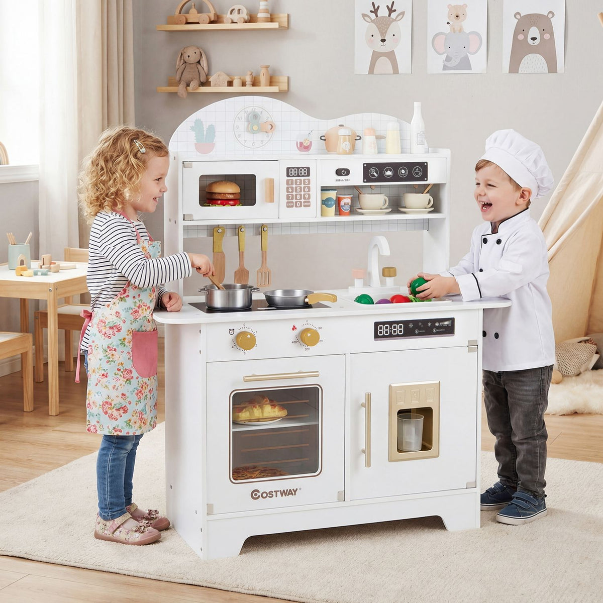 Two children play at a white toy kitchen, with pots on the stovetop and utensils hanging above.