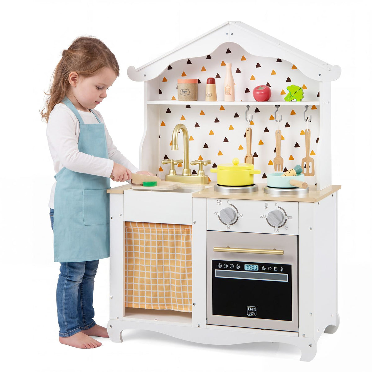 A child in an apron plays at the white kitchen, using a wooden utensil beside the toy sink and stove.