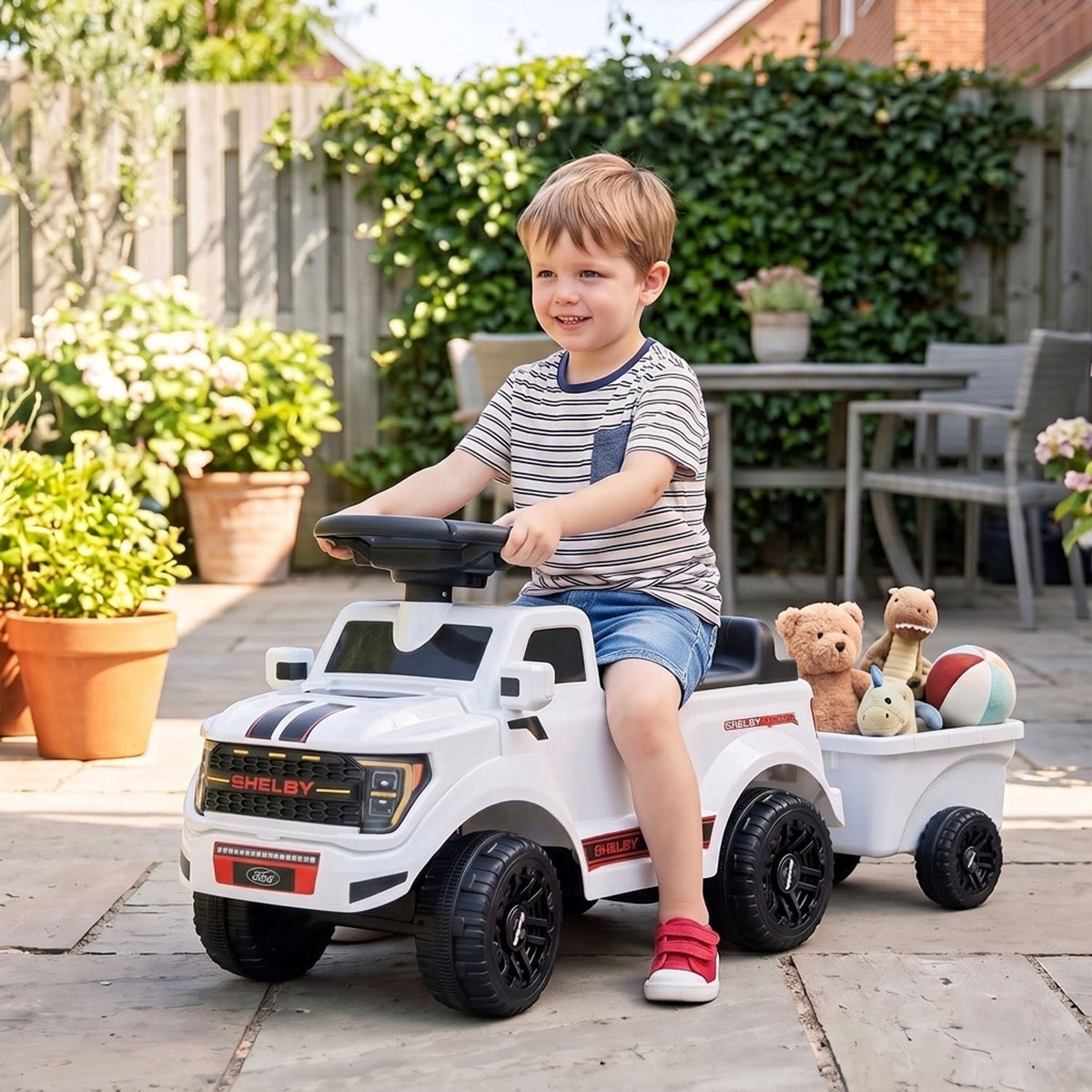A child drives a white ride-on car towing a trailer with stuffed toys in a garden.