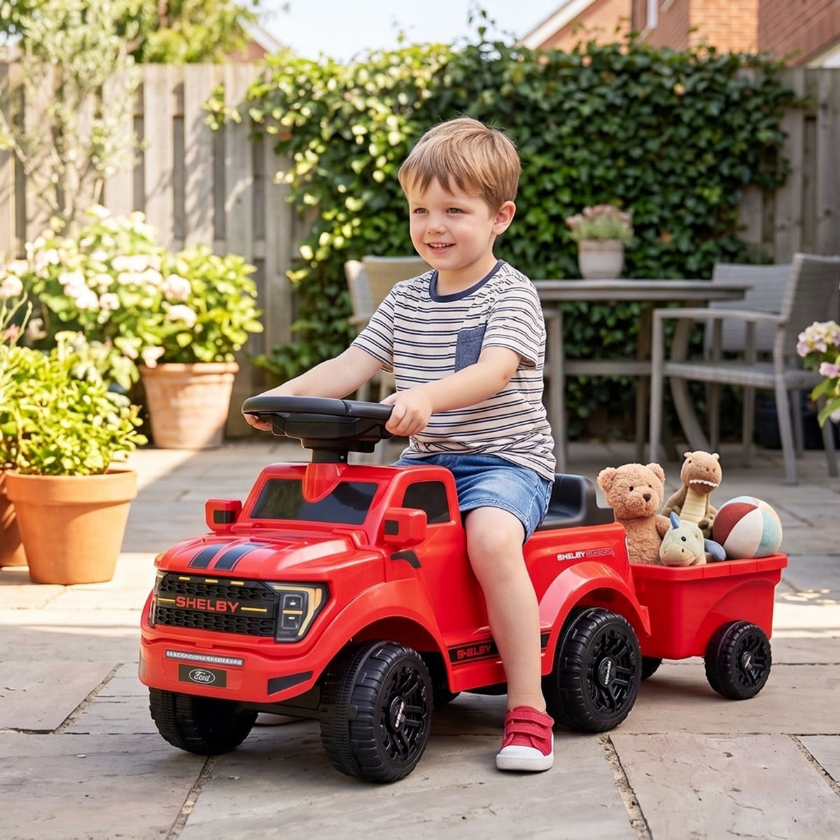 A child sits in a red ride-on truck towing a trailer with stuffed toys in a sunny patio garden.
