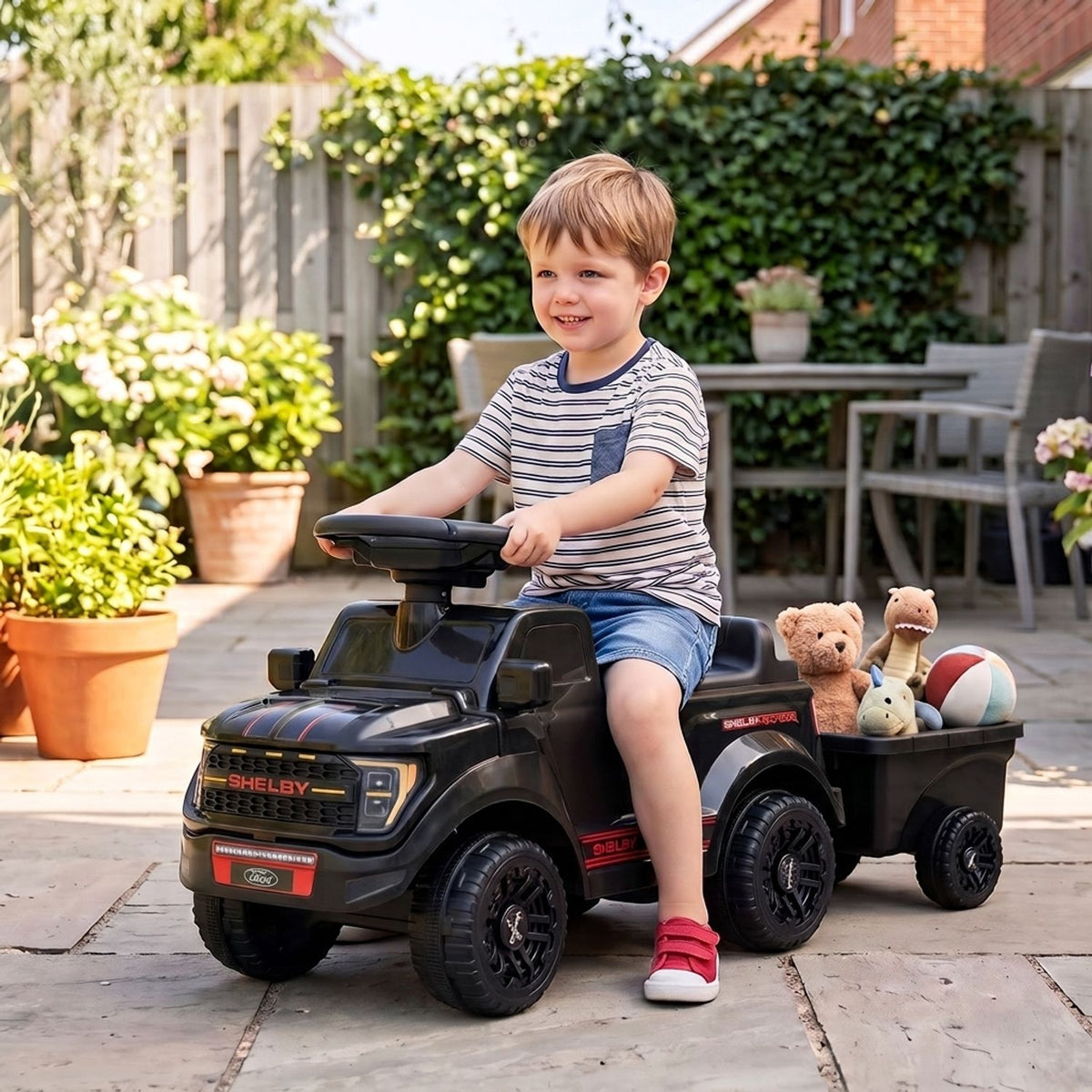 A child sits on a black ride-on car with a trailer carrying toys in a garden patio.