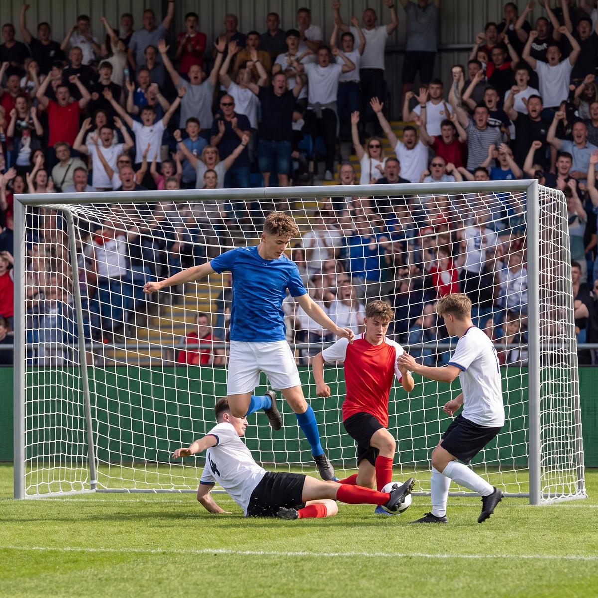 Players contest the ball in front of a soccer goal as spectators cheer in the stands.
