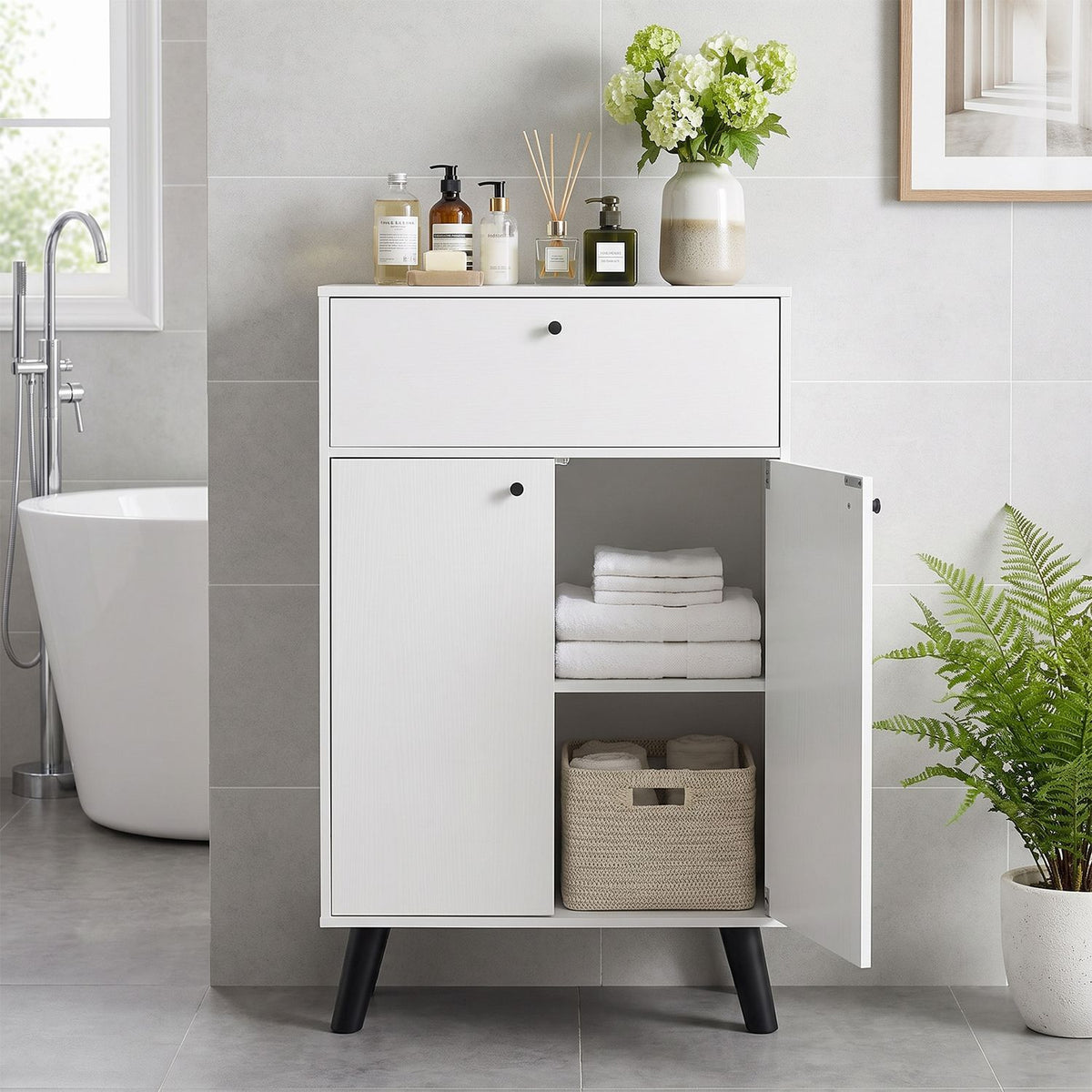 White cabinet with a drawer and open lower door displaying folded towels and a woven basket in a tiled bathroom.