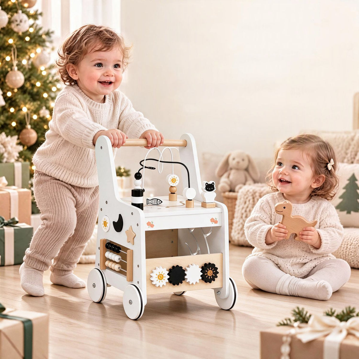 Two toddlers play beside a white activity walker in a softly lit holiday-decorated room.