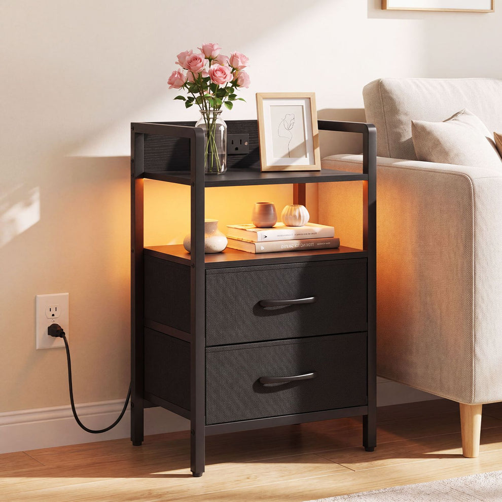 Black nightstand beside a sofa, with two fabric drawers, an open shelf glowing amber, and flowers on top.