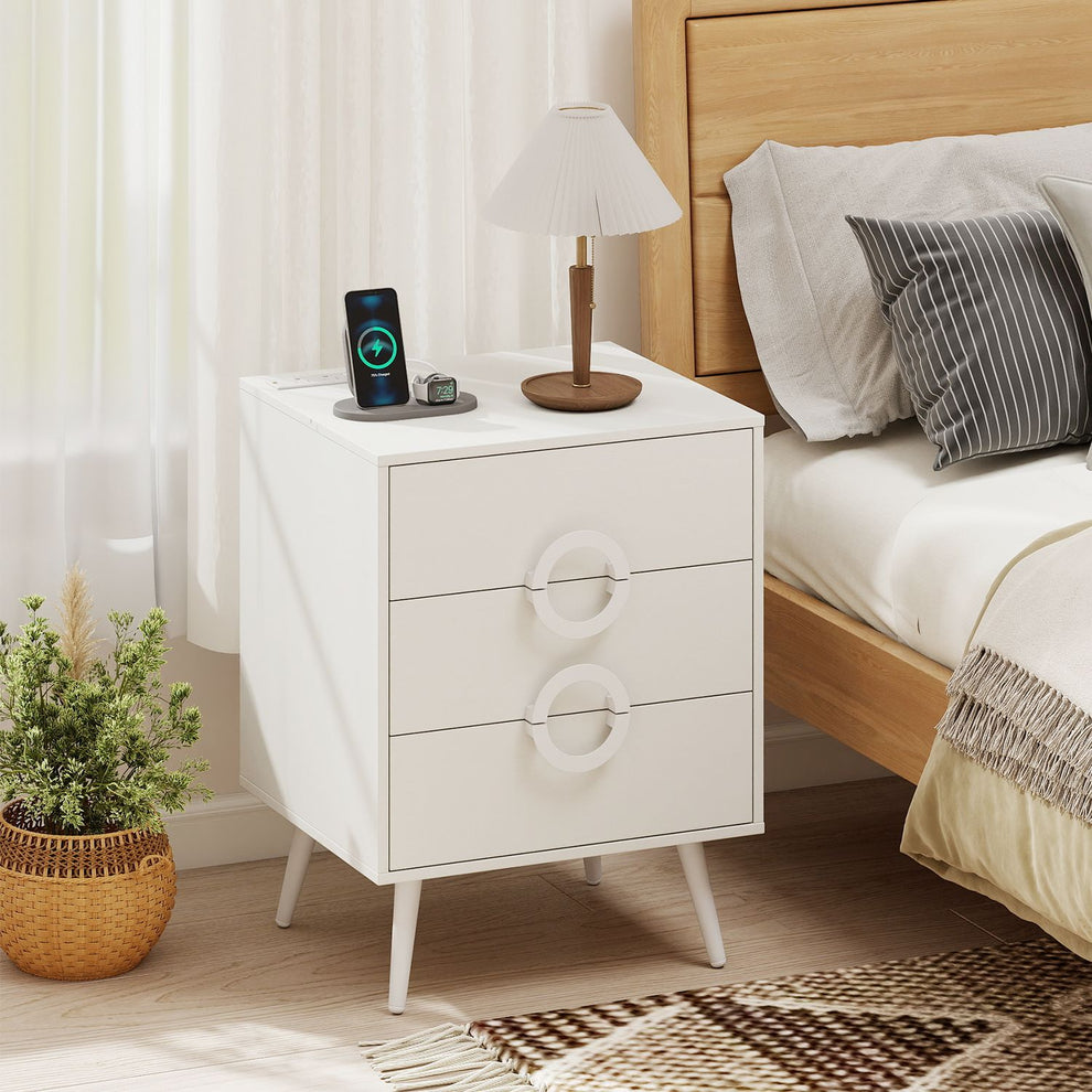 White bedside nightstand with circular drawer pulls beside a bed, angled front view