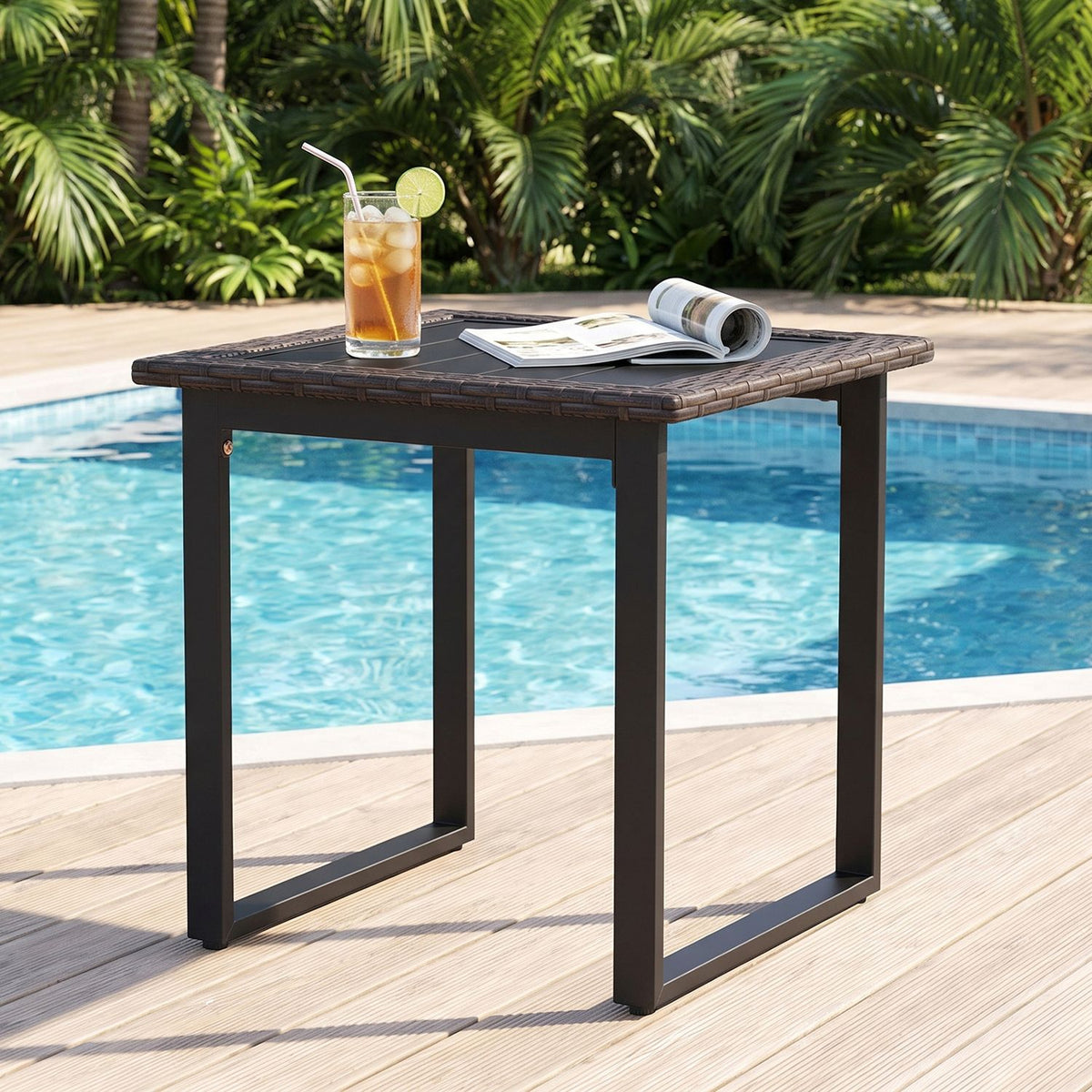 A dark square side table beside a swimming pool, holding an iced drink and an open magazine.