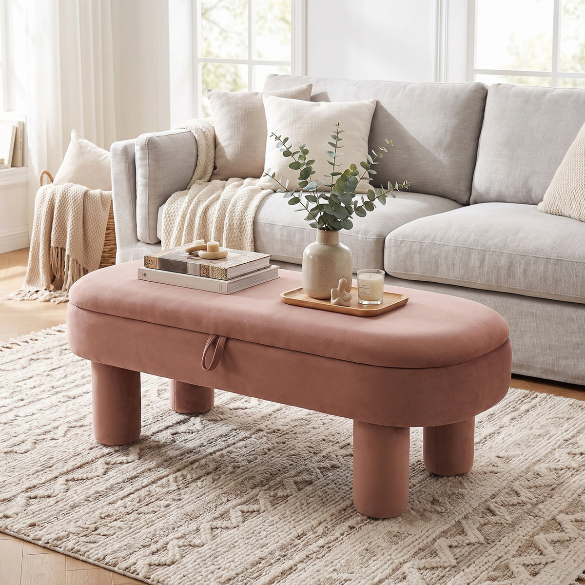 Dusty rose oval ottoman bench in front of a sofa, styled with a tray and vase.
