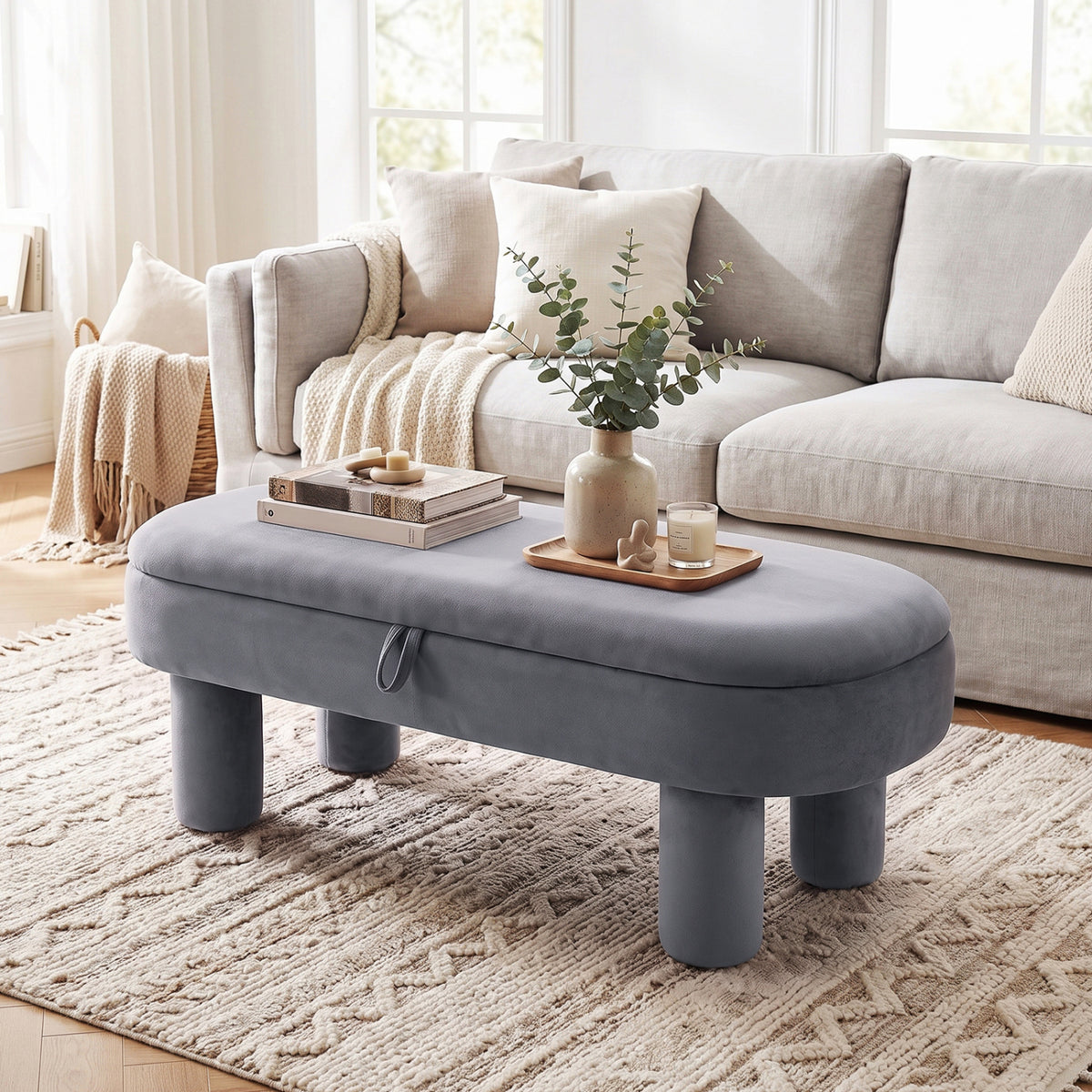 Gray oval ottoman bench in front of a light sofa, styled with books and a tray on a rug.
