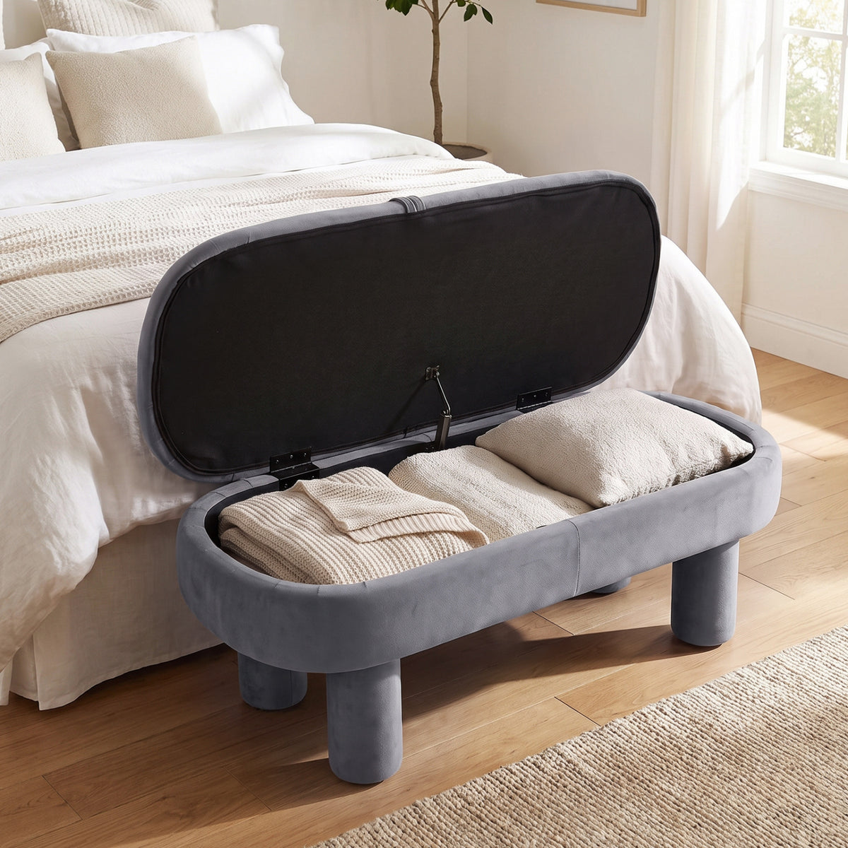 Gray upholstered bench opened beside a bed, with folded blankets and pillows inside.