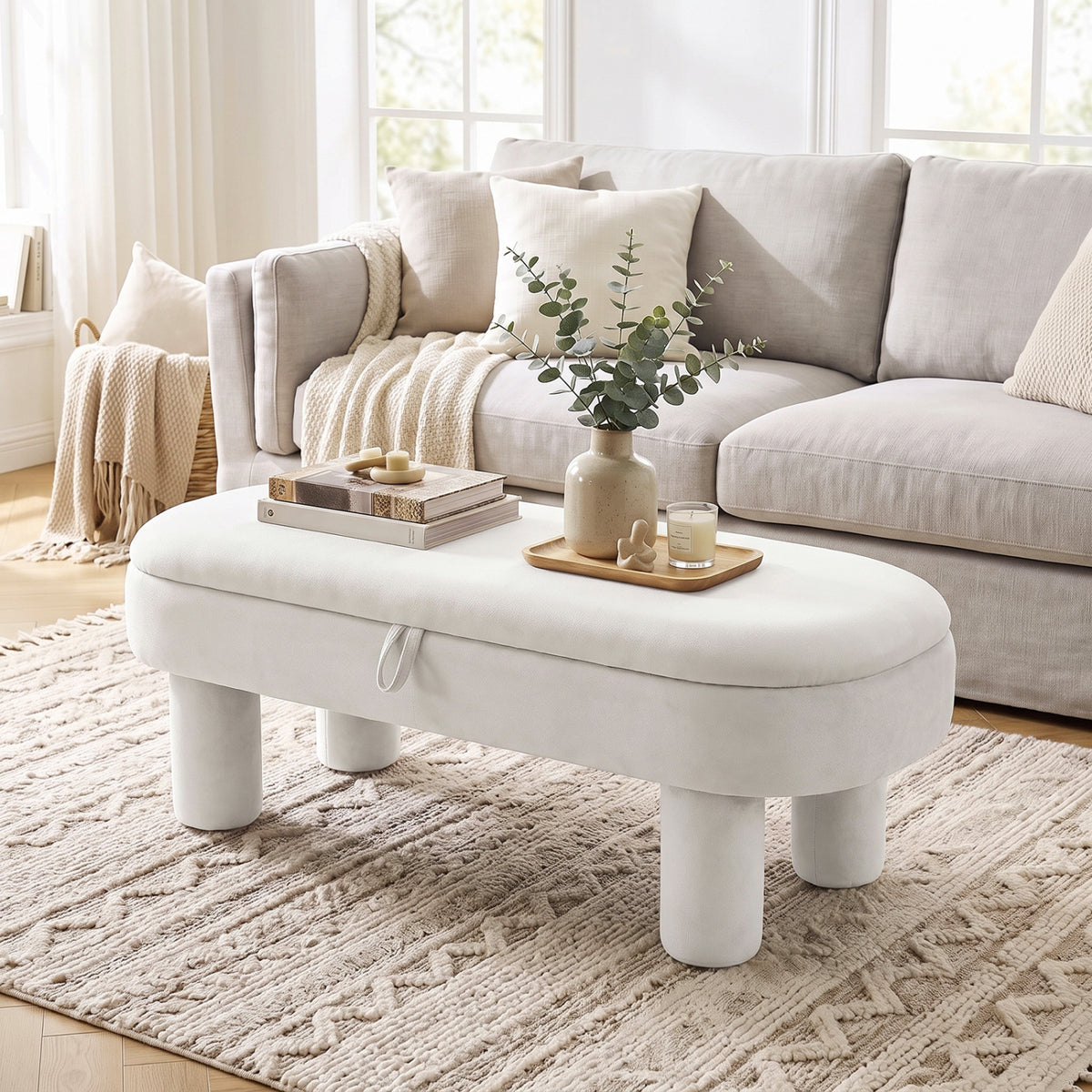Cream oval ottoman bench with a cushioned top and rounded legs in a bright living room.