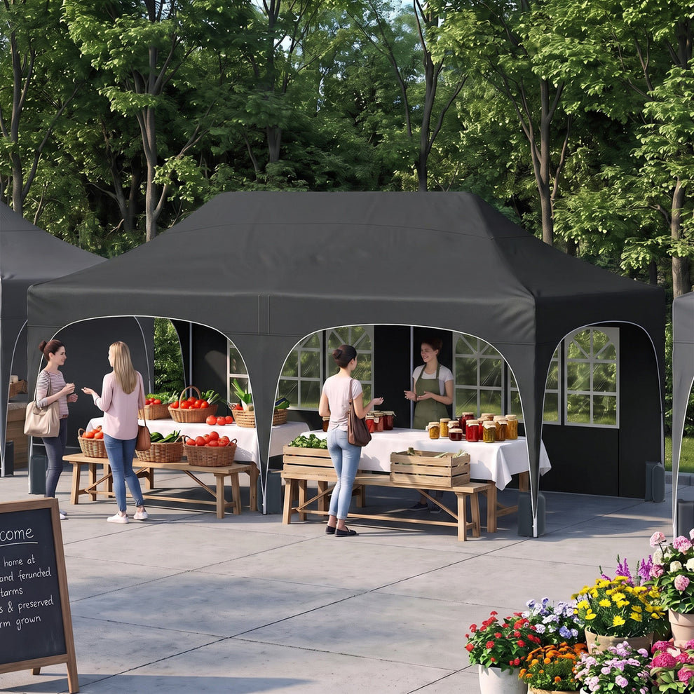 A dark gray canopy tent with arched open sides shelters market tables and produce in a garden setting.