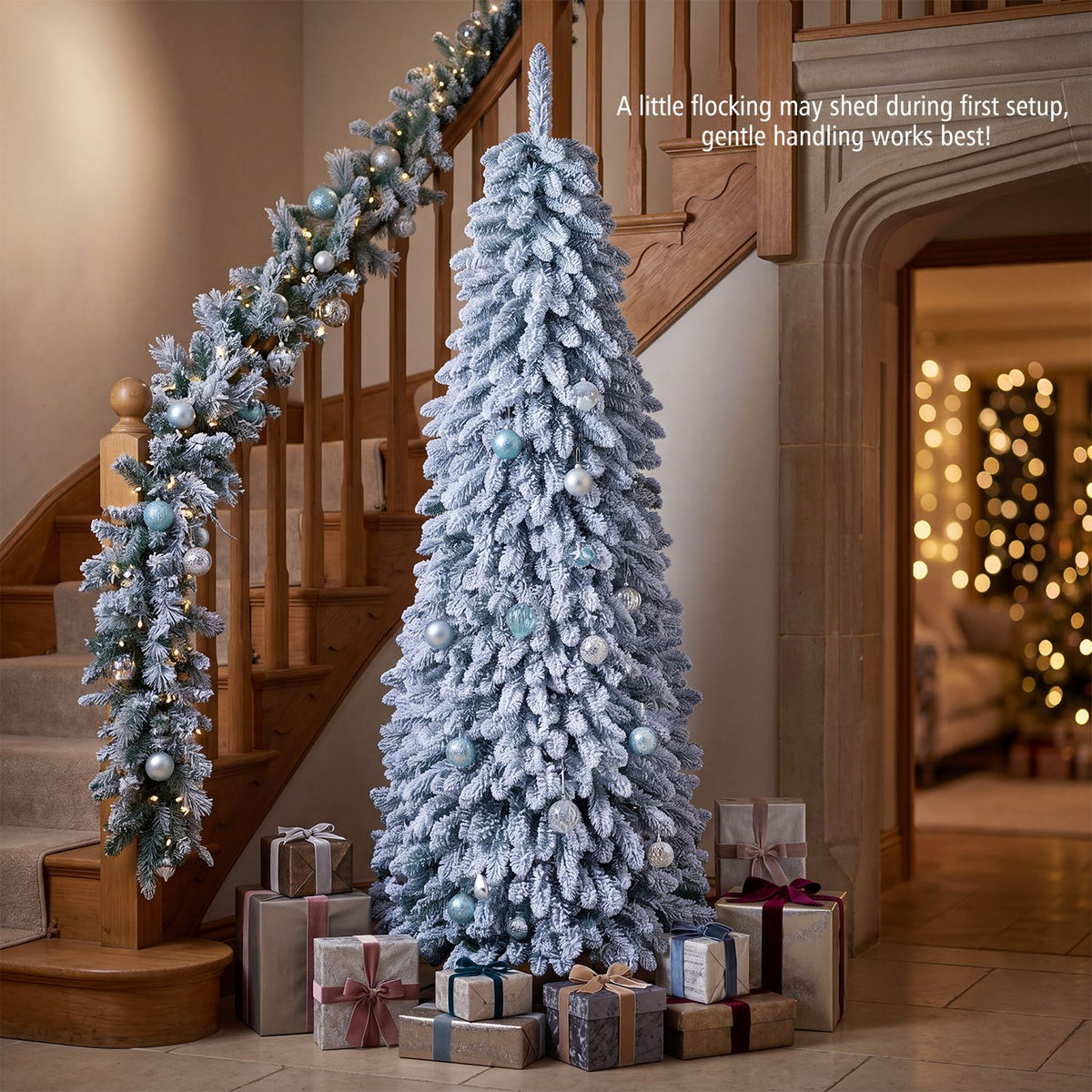Snow-flocked Christmas tree decorated with silver and blue ornaments beside a staircase, with wrapped gifts below.