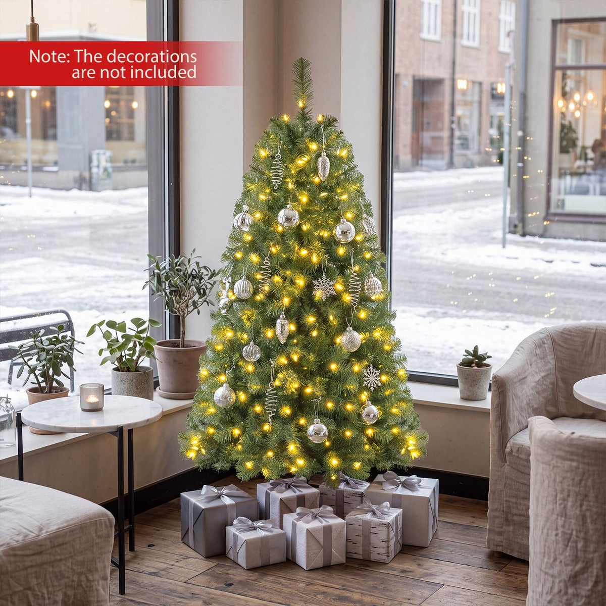 A lit Christmas tree with silver decorations and wrapped gifts stands by large windows.