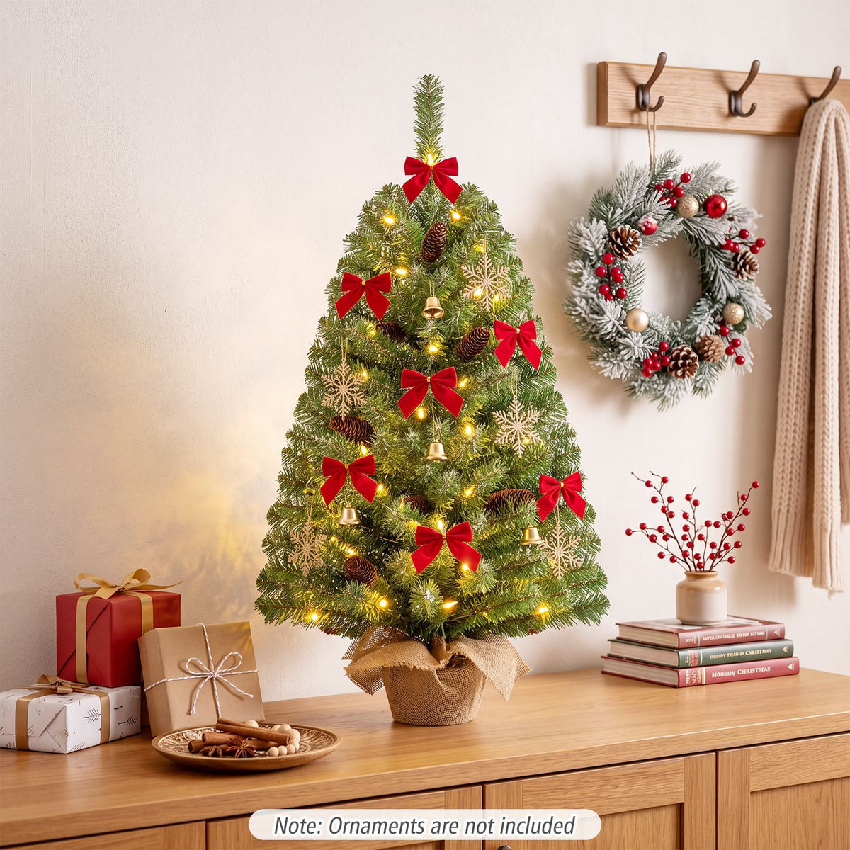 A lit tabletop tree decorated with red bows and gold ornaments sits on a wooden table beside a wreath.