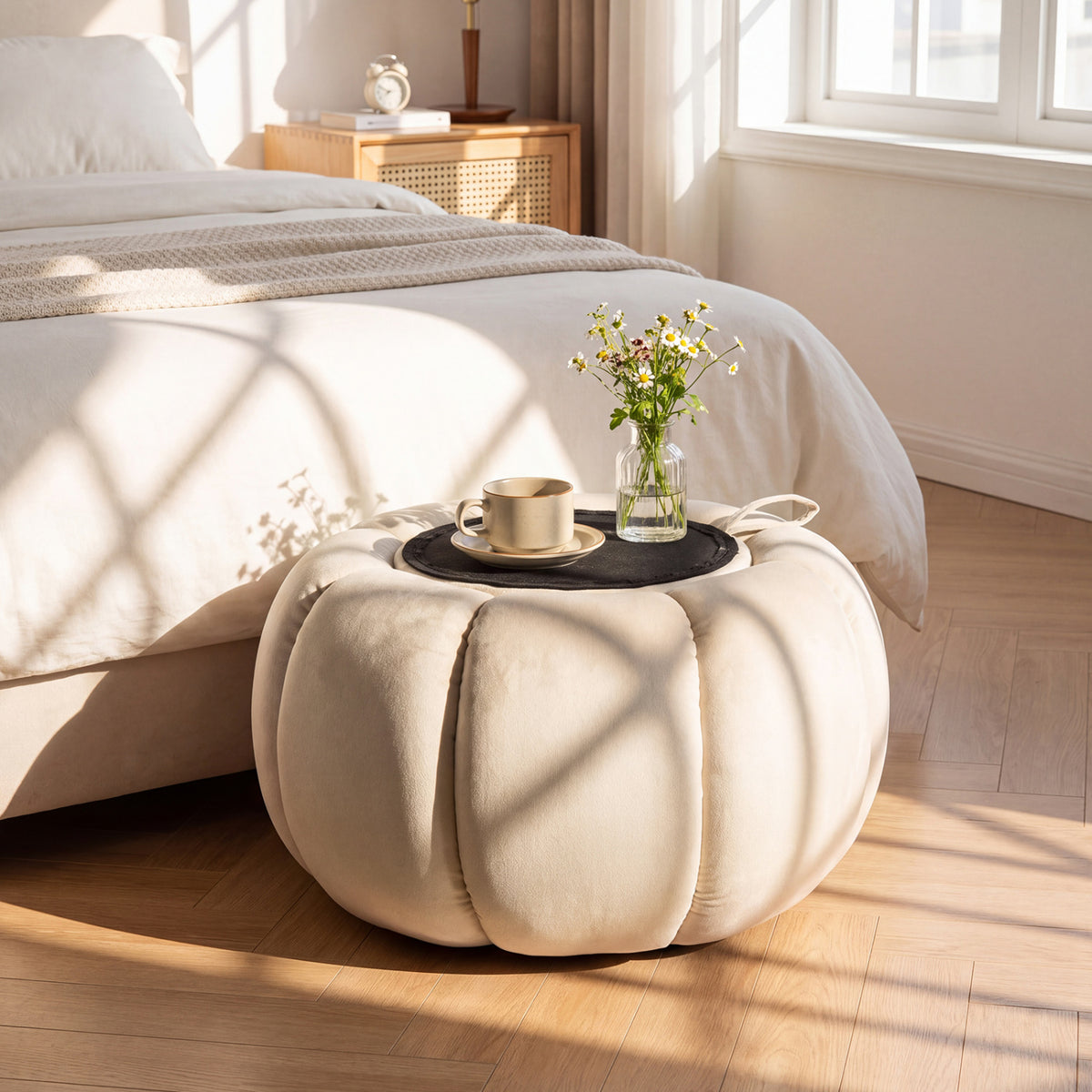 Round cream ottoman beside a bed, topped with a tray, a cup, and a vase of flowers.