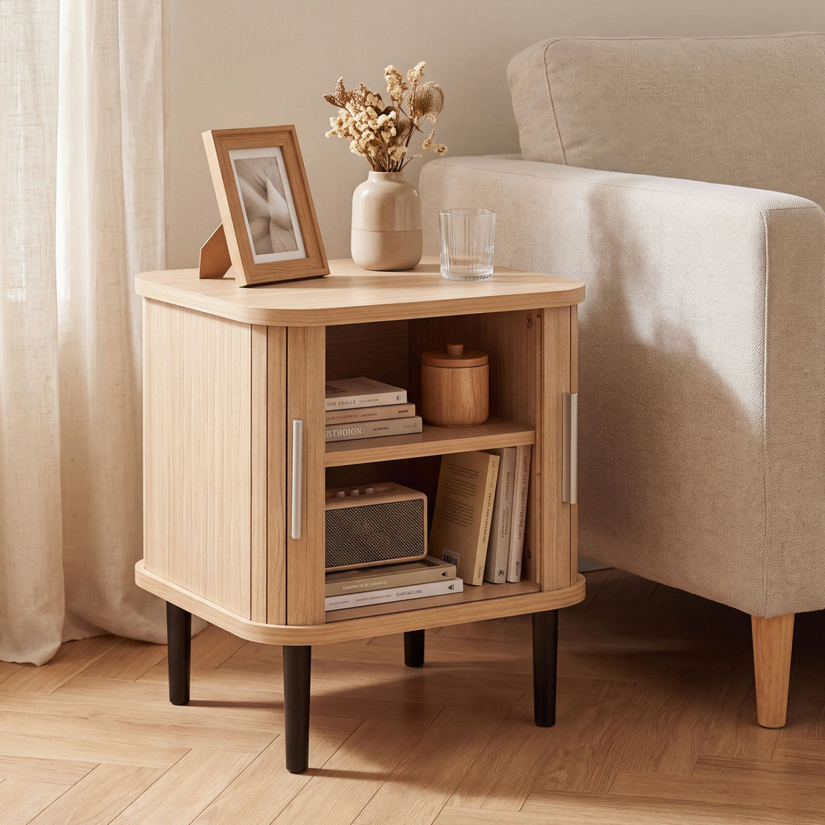 Fluted light-wood nightstand beside an upholstered chair, with its sliding doors open to books and decor.