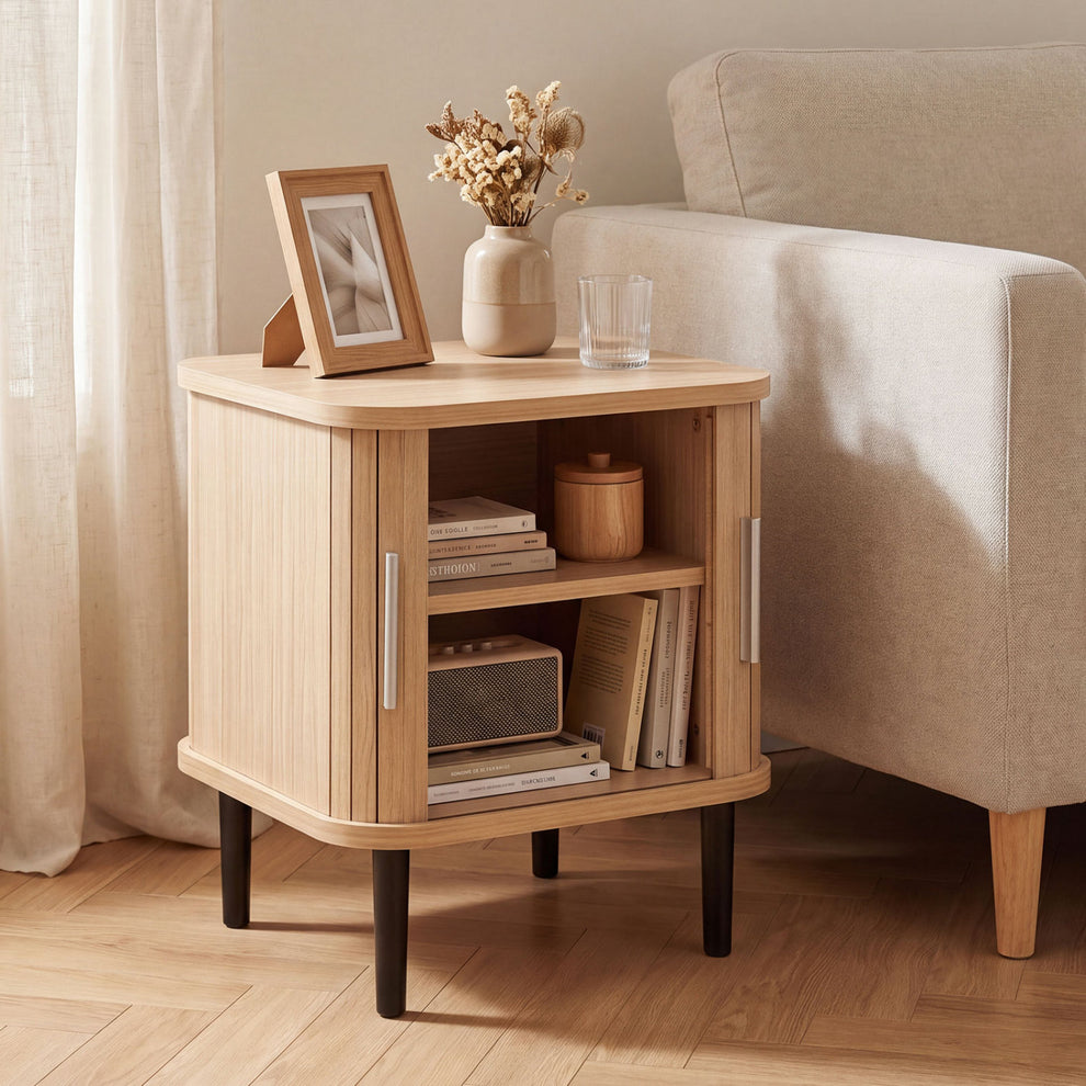 Fluted light-wood nightstand beside an upholstered chair, with its sliding doors open to books and decor.