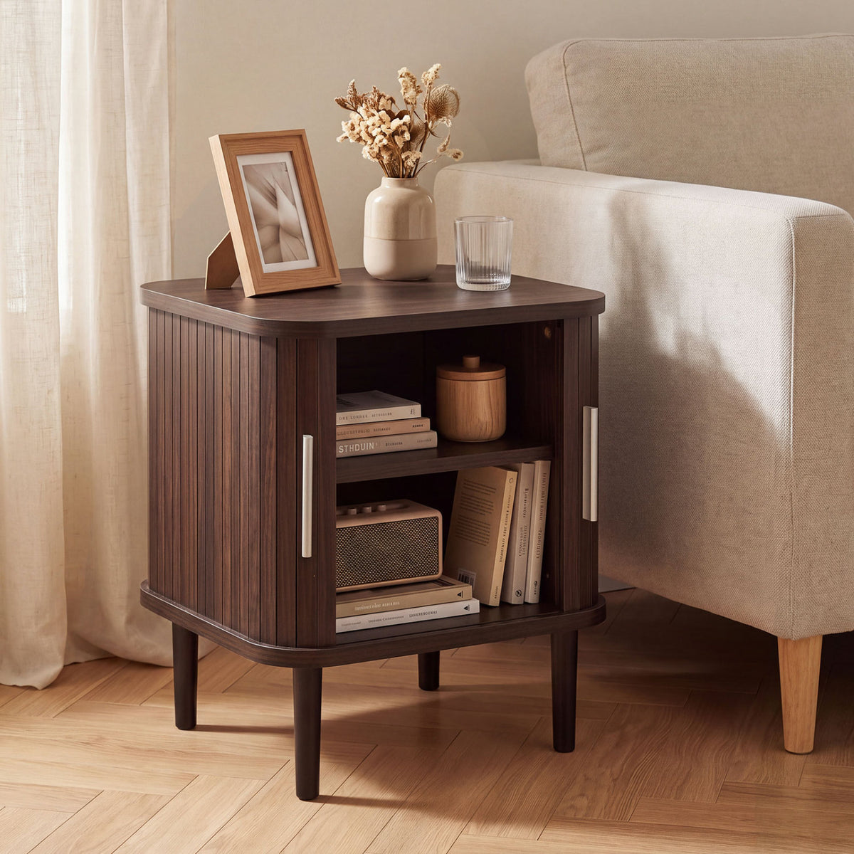 Dark wood fluted nightstand with sliding doors beside a beige chair, styled with books and decor.