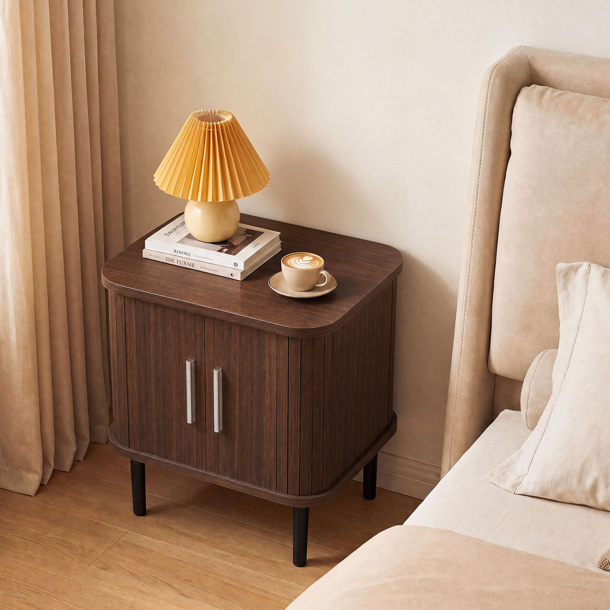 Closed dark fluted nightstand beside a cream bed, topped with a pleated lamp, books, and a cup.