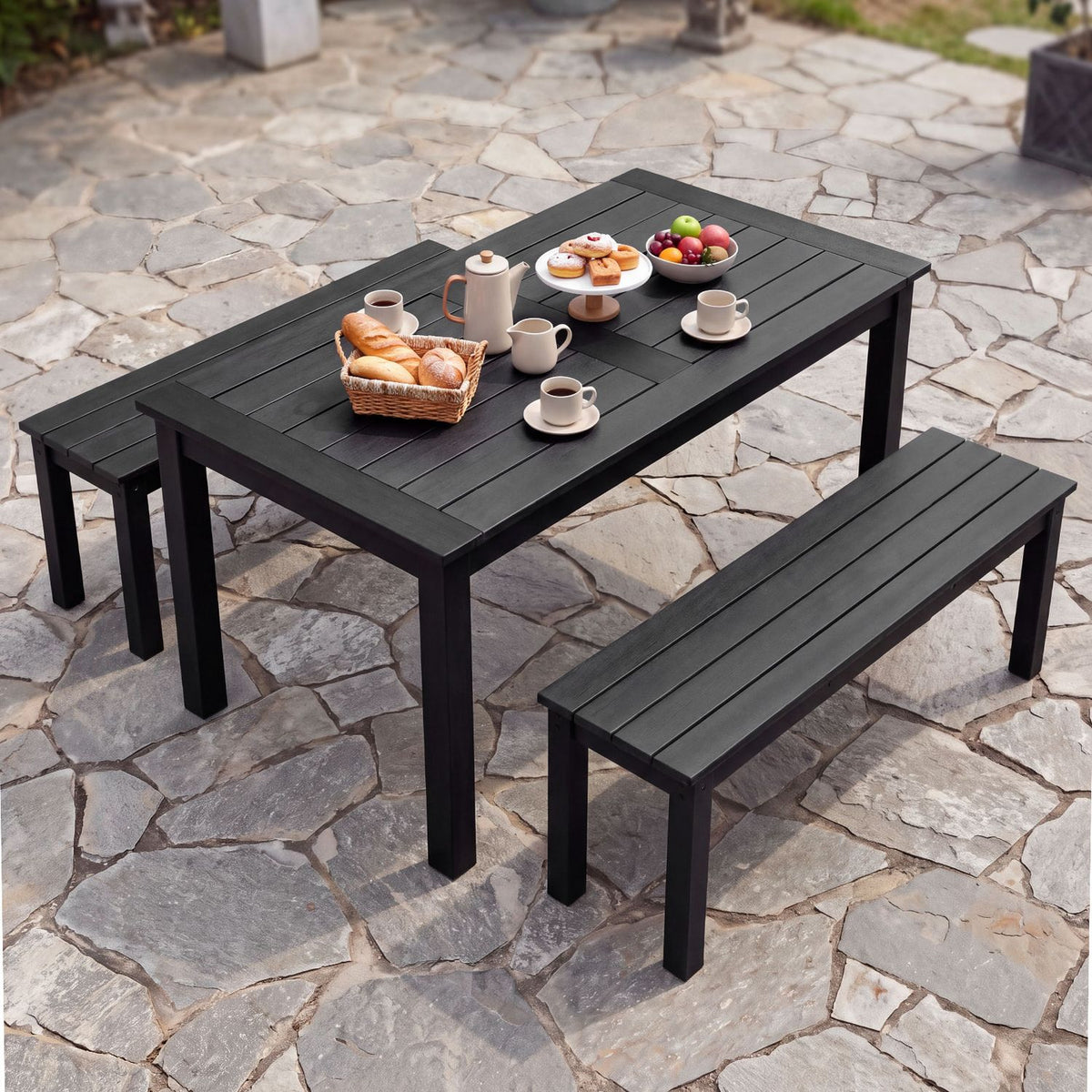 Two dark slatted benches sit beside a matching outdoor table set with cups, bread, and fruit on a stone patio.