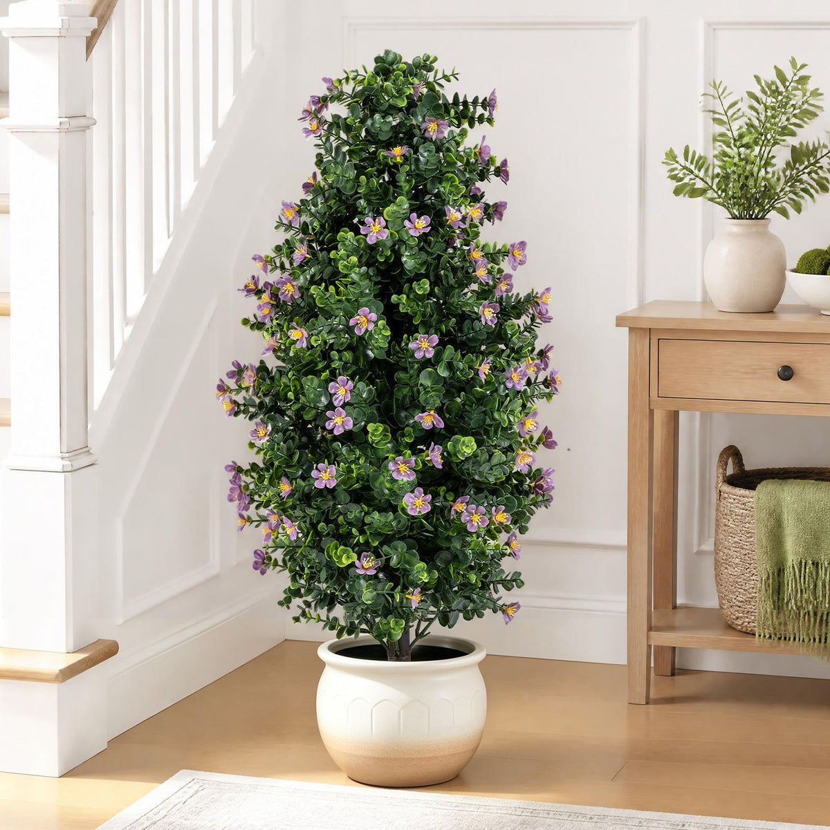 Purple-blossomed artificial topiary in a white planter beside a wooden console and staircase.