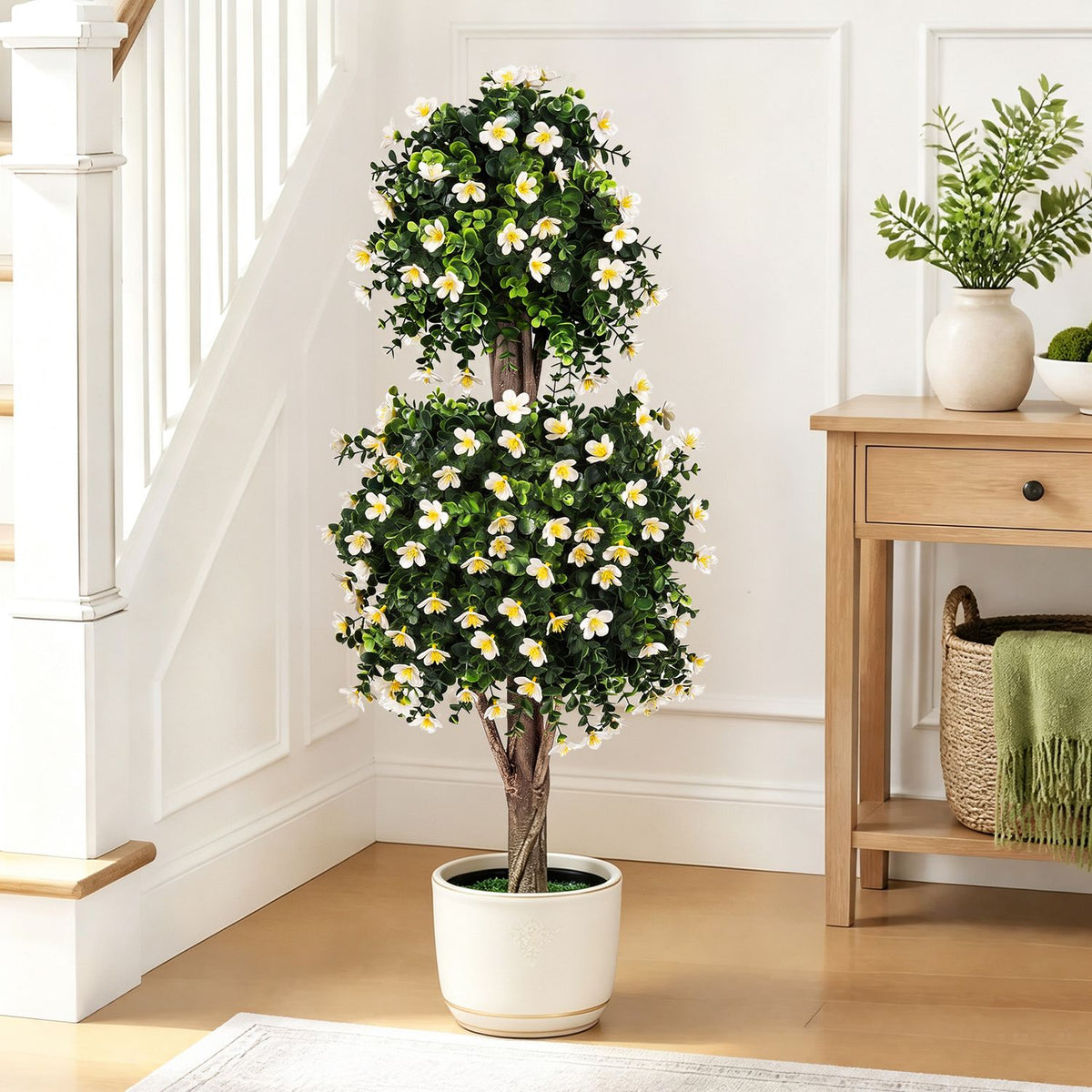 White-flowered two-ball topiary tree in a white pot beside a staircase and wooden console.