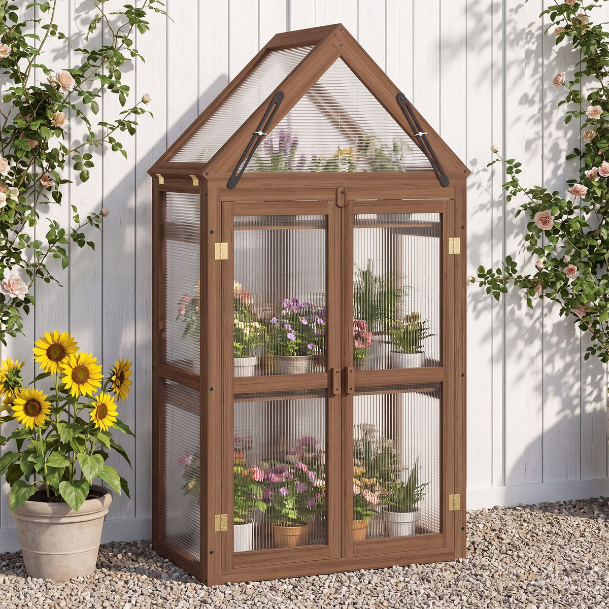 Front view of a wood-framed greenhouse with closed double doors and plants inside, set against a white fence.