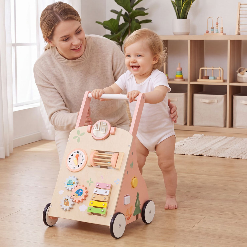 A smiling toddler pushes a wooden activity walker while an adult supports her in a bright room.