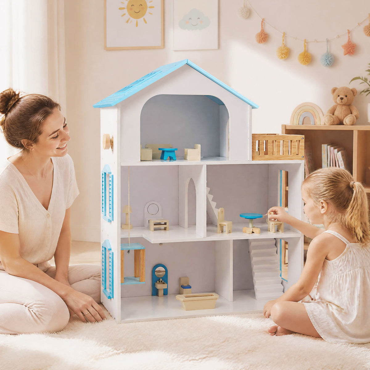 A woman and child play beside an open white dollhouse with blue trim and miniature furniture.
