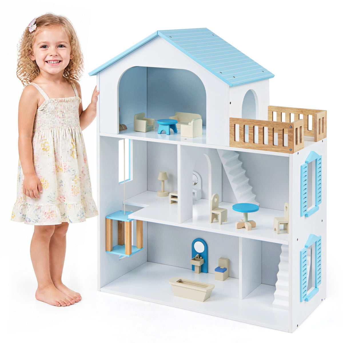 A child stands beside a white dollhouse with blue roof trim, wooden balcony railings and miniature furniture.