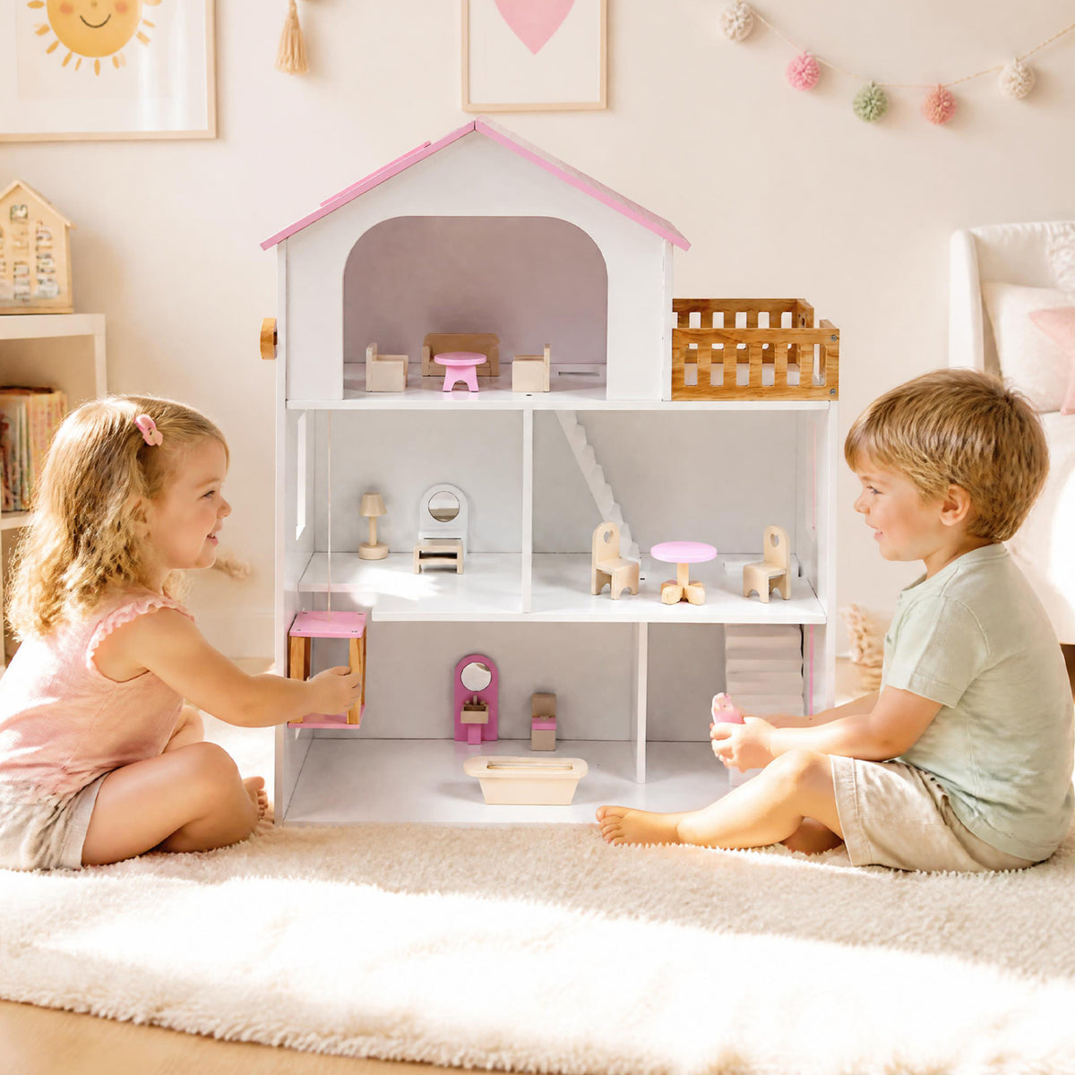 Two children sit beside a multi-level dollhouse with miniature furniture and a wooden balcony.