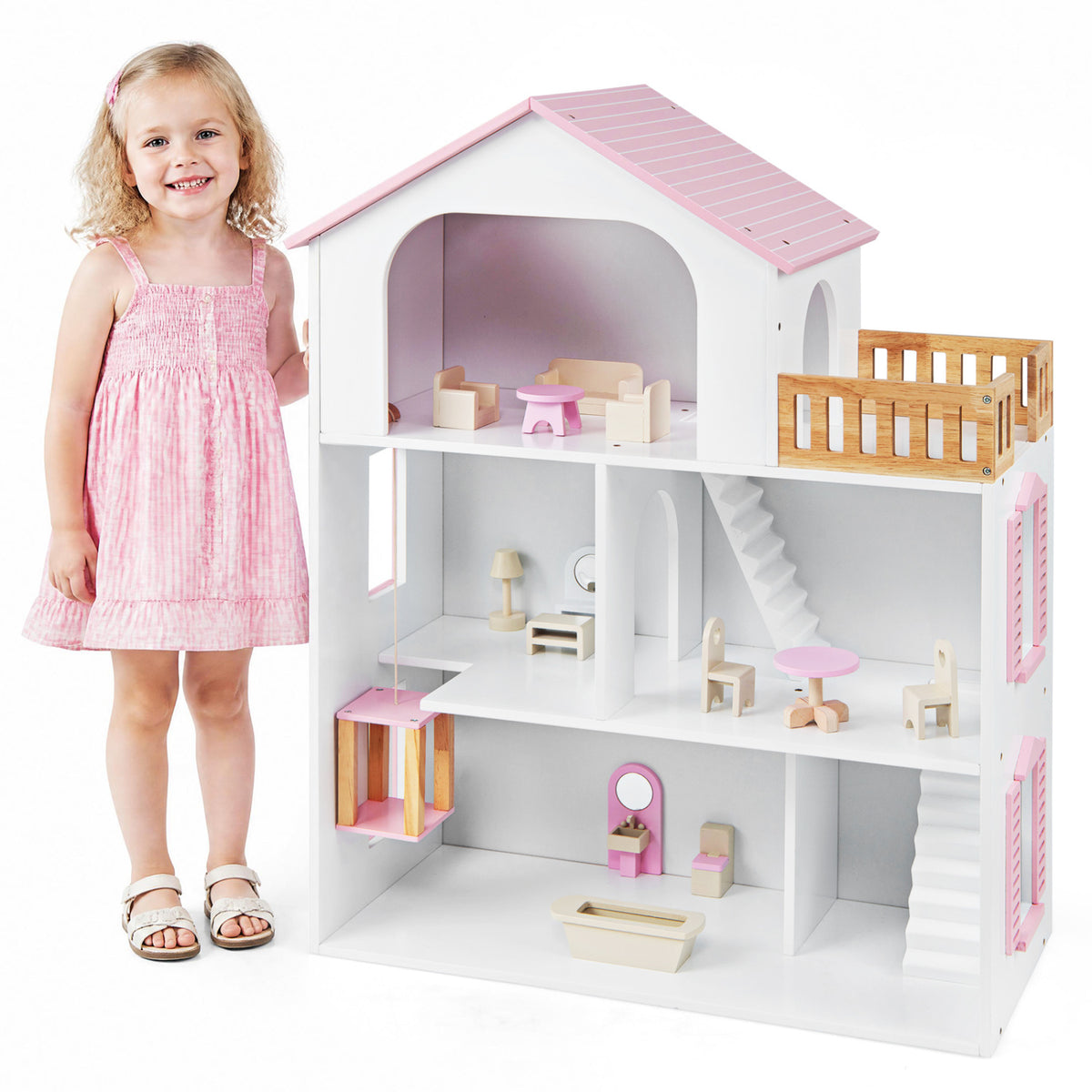 A girl stands beside the open-front dollhouse, showing its pink roof, wooden balcony, stairs, and furnished rooms.