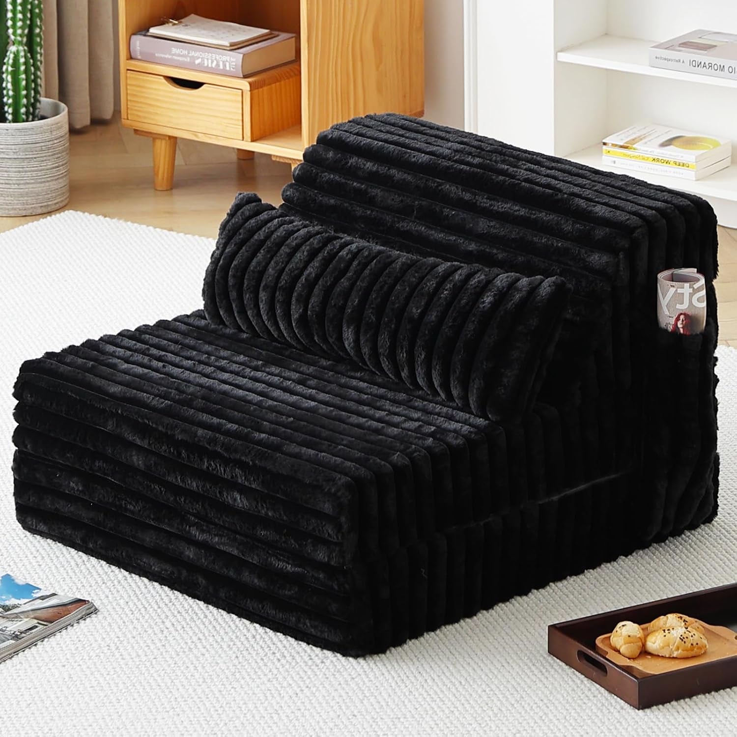 Black ribbed folding sofa bed arranged as a compact seat with cylindrical cushion and magazine in the side pocket.