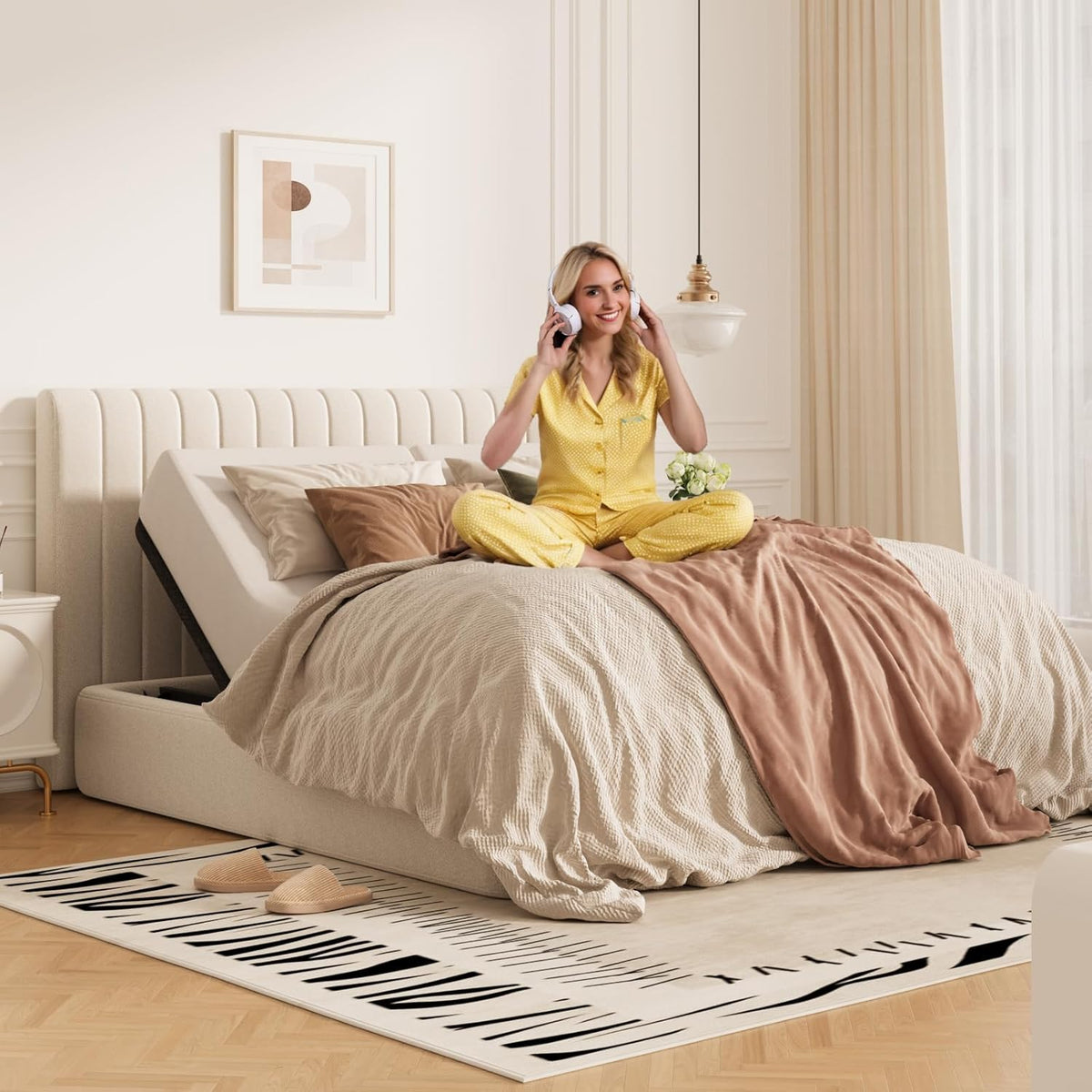 Front three-quarter bedroom view: beige upholstered adjustable base with head raised, woman wearing headphones seated