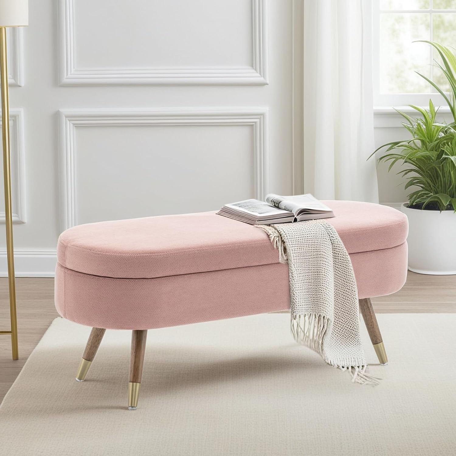 Oval pink upholstered bench with wooden legs and gold caps shown with a throw and open book.