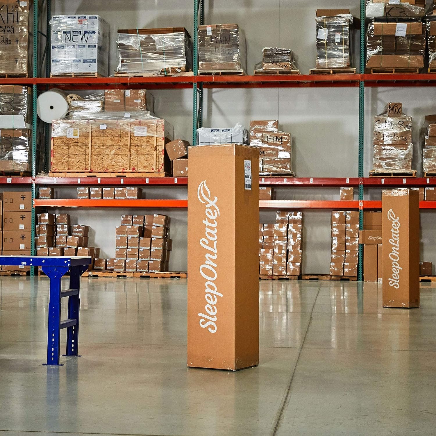 Tall boxed mattress standing upright in a warehouse among shelving and packaged inventory.
