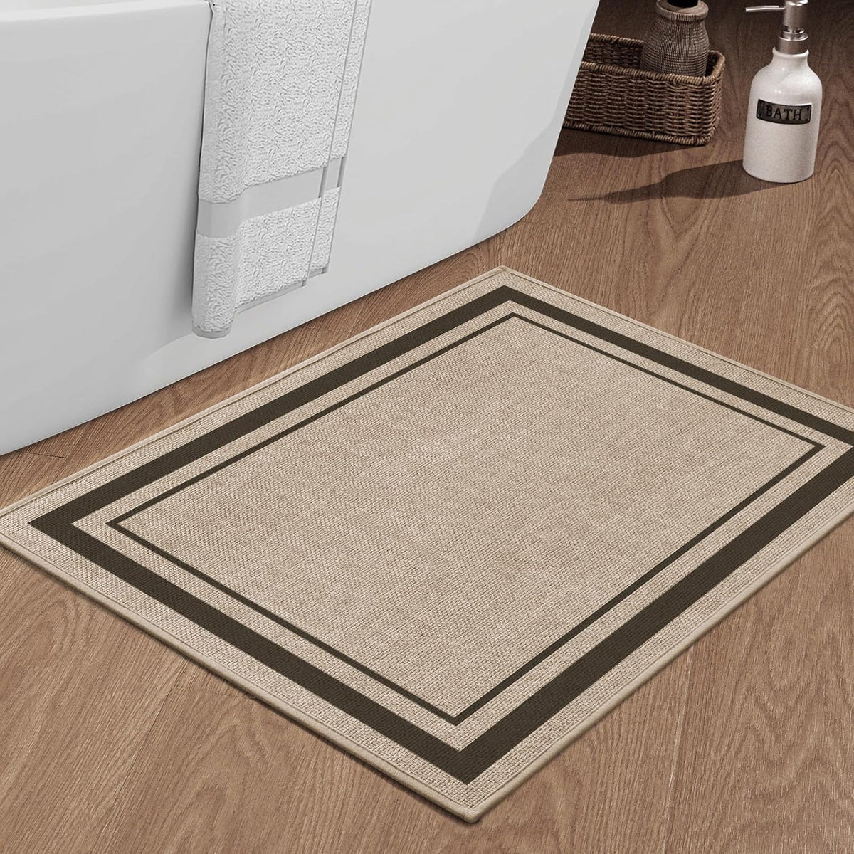 Small rectangular beige bath mat with dark border set on wood floor beside a bathtub and towel.