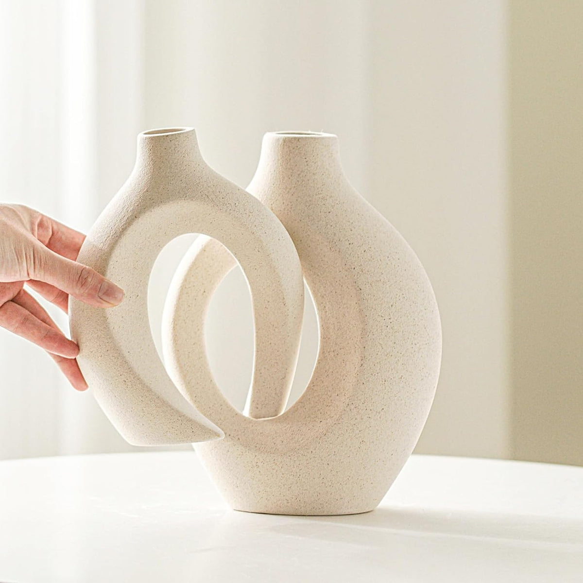 Hand holding the smaller matte beige hollow vase next to the larger vase in a close-up studio view