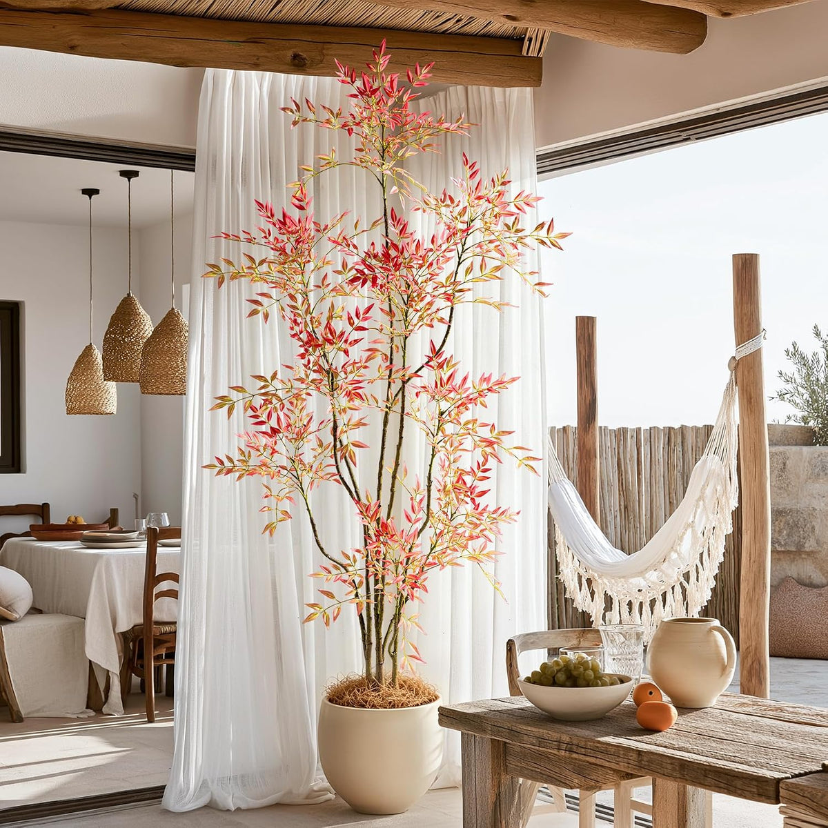 Tall faux Nandina with red and gold leaves in a round beige planter beside a hammock and dining table