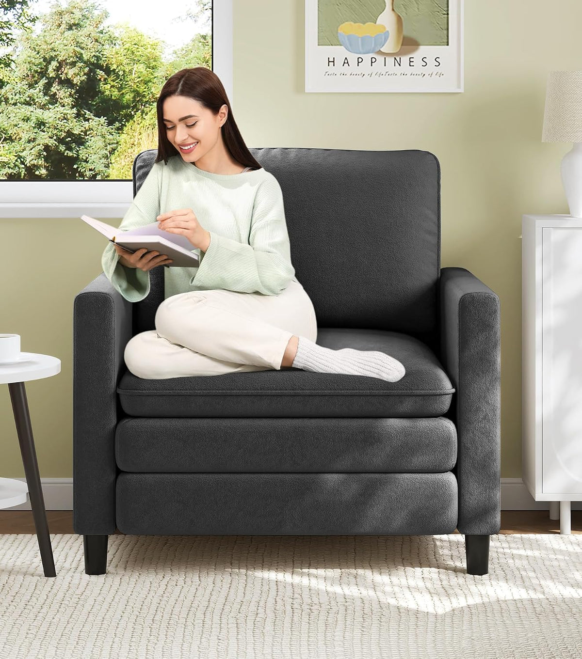 Woman sitting cross-legged reading on a charcoal-gray small loveseat in a sunlit room.