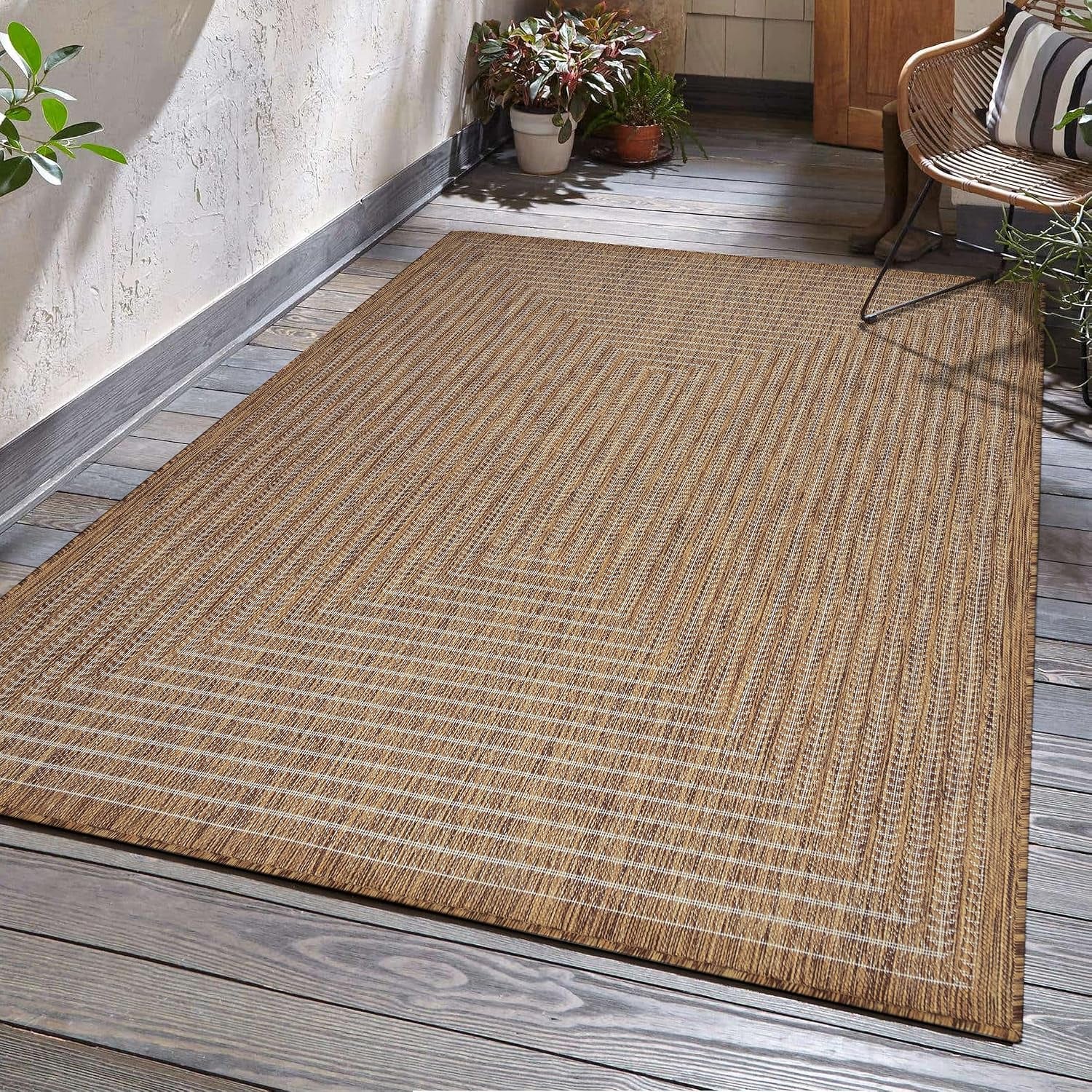 Natural beige geometric-border rug on an outdoor deck with a rattan chair and potted plants at upper right