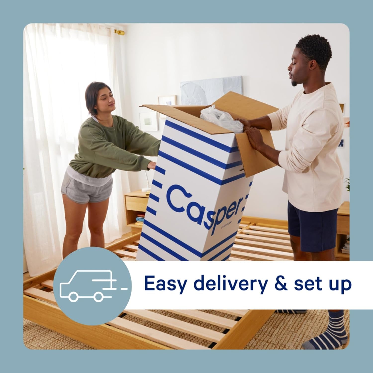 Two people unpacking a tall striped mattress shipping box at the foot of a wooden slat bed frame.