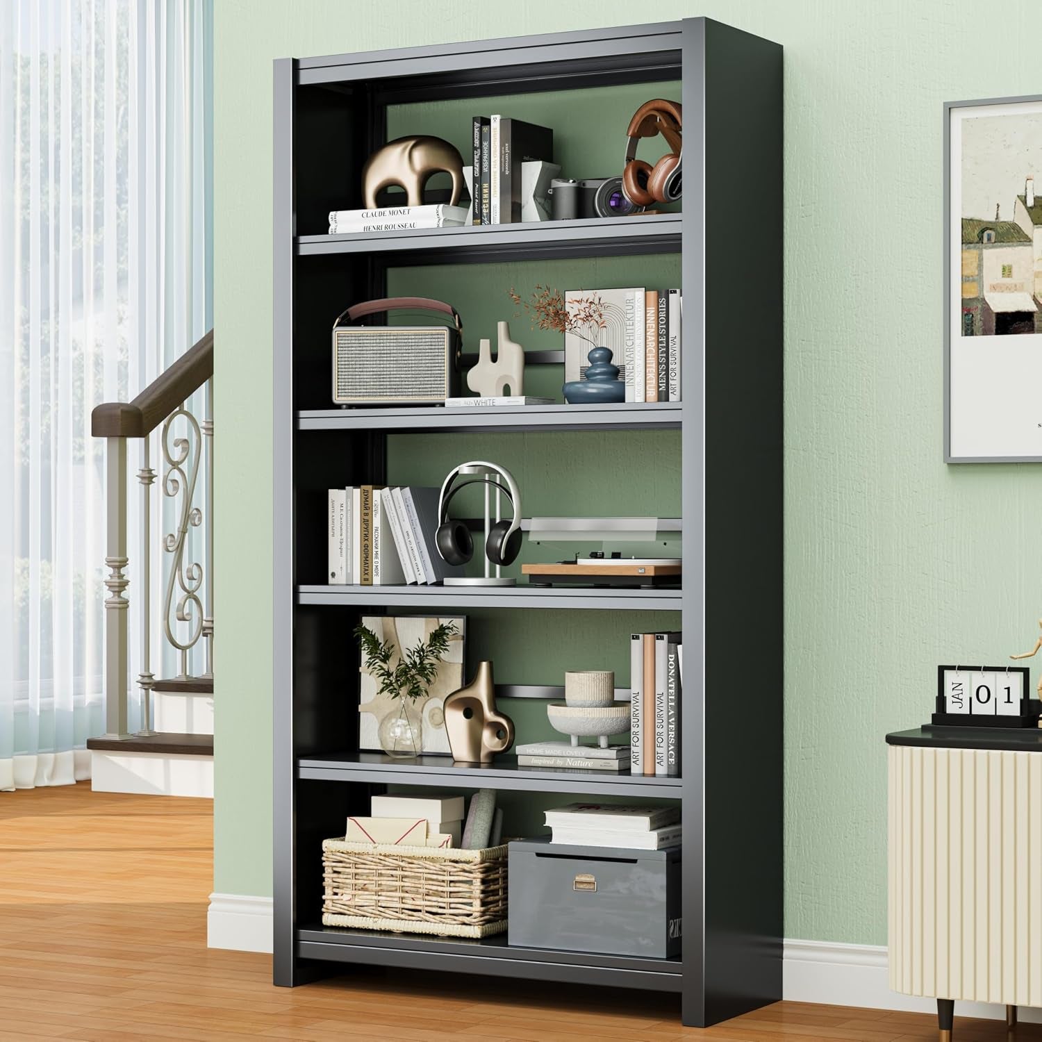 Tall black metal shelving unit against a green wall, styled with books, headphones, basket and decorative objects; staircase