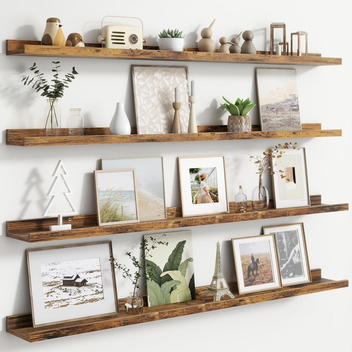 Three long rustic wood floating picture ledges mounted on a white wall holding framed art, plants, and decor.